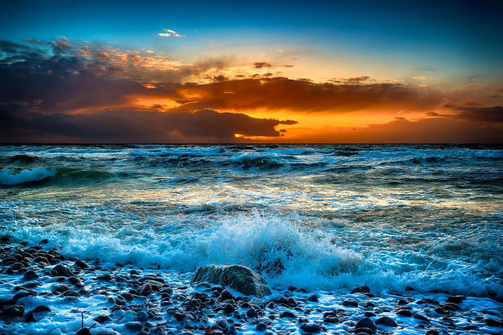 Wellen brechen an einem steinigen Strand bei Sonnenuntergang mit dramatischem Himmel.