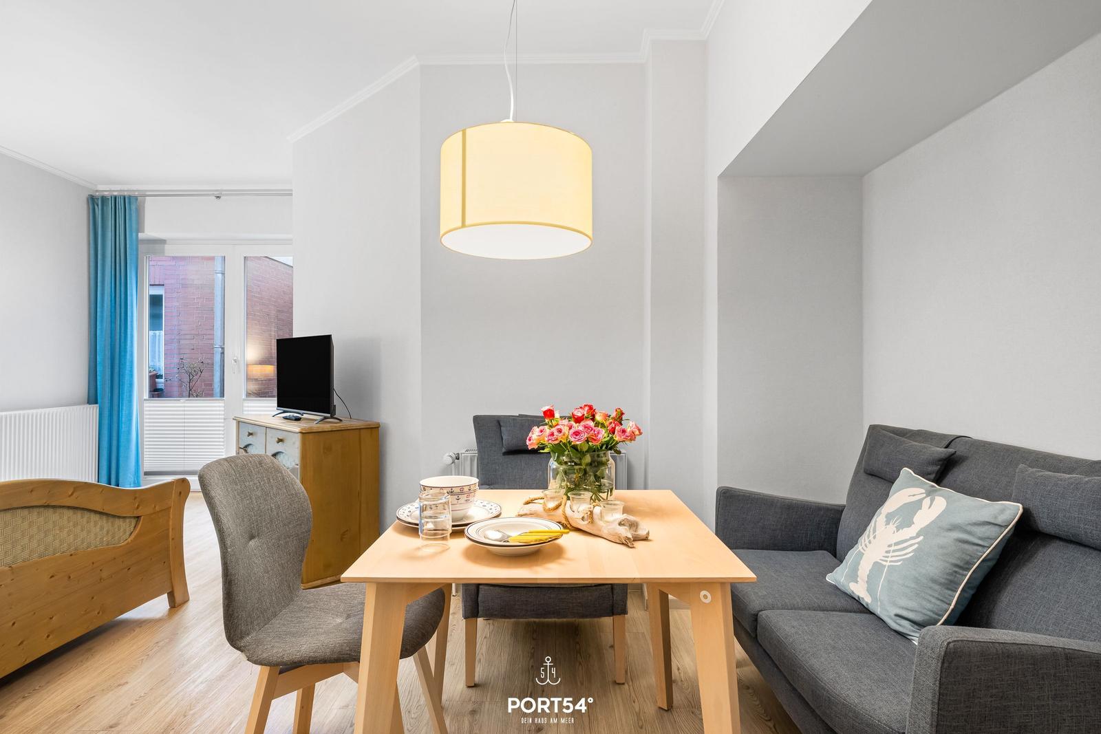 Esszimmer mit Tisch, Stühlen und Sofa. Blumenvase und TV sind sichtbar.