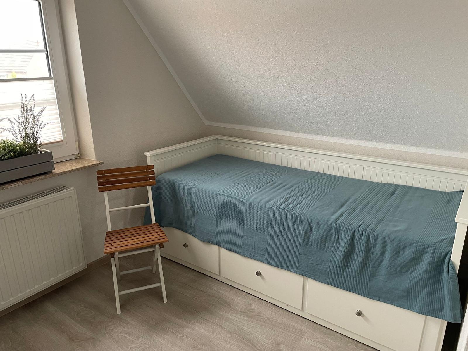 Ein Schlafzimmer mit Dachschrägen, weißem Bett und Holzstuhl neben dem Fenster.