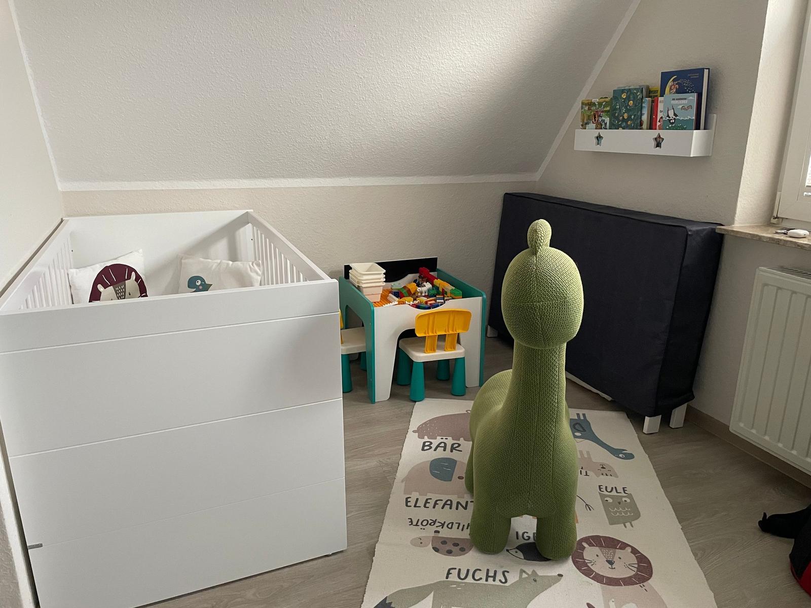 Kinderzimmer mit Babybett, Spielzeugtisch und grünem Stofftier auf Tiermuster-Teppich.