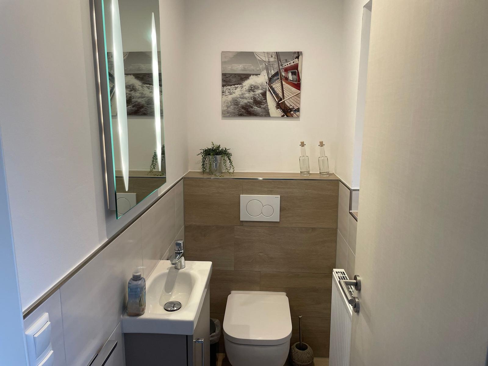 Badezimmer mit Waschbecken, Toilette und Spiegel. Wandbild zeigt Segelyacht.