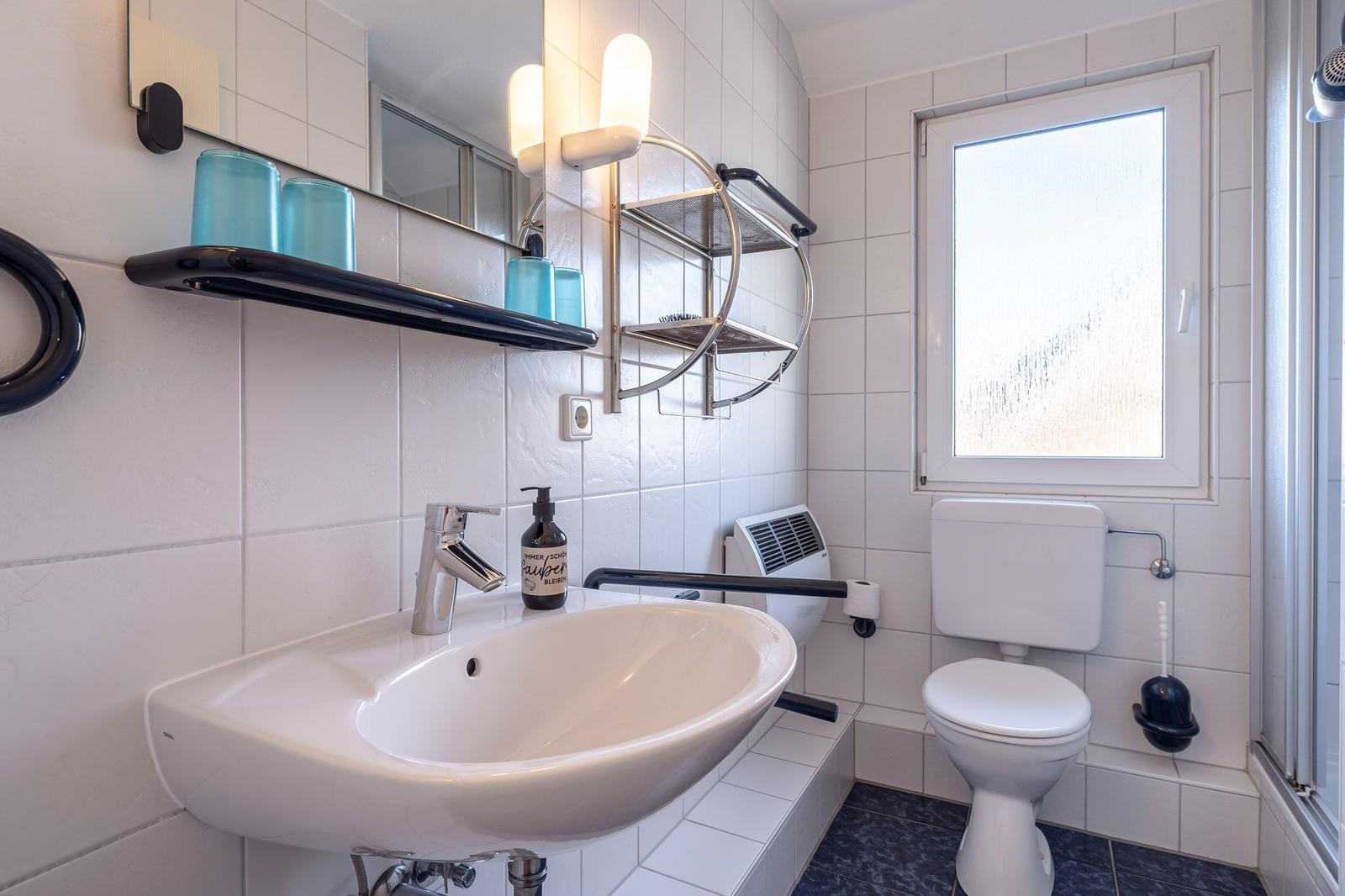 Badezimmer mit Waschbecken, Toilette und Fenster. Weiße Fliesen und blaue Accessoires.