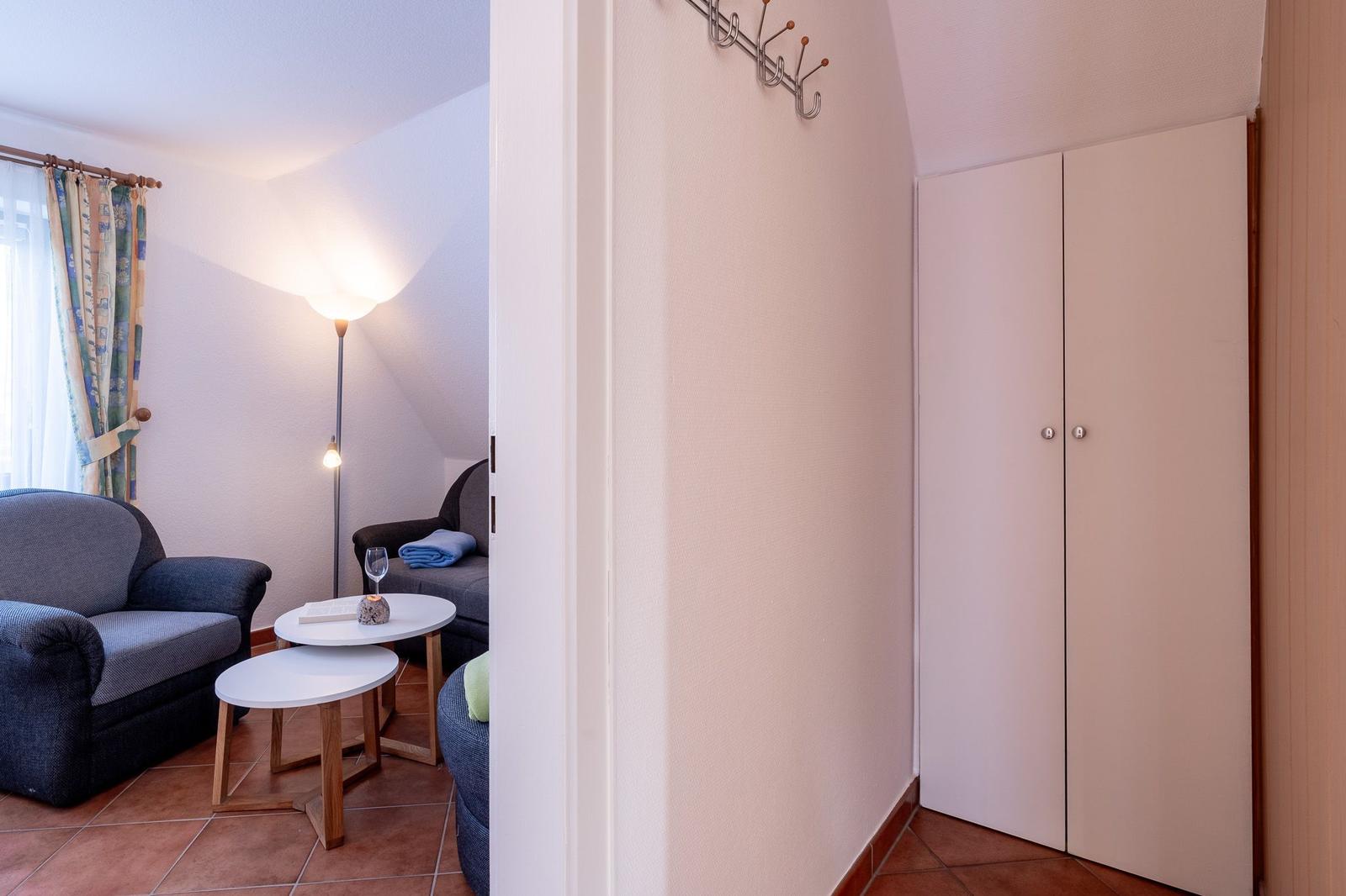 Wohnbereich mit Sitzgelegenheit, Tischen und Gardinen. Flur mit Kleiderständer und Schrank.