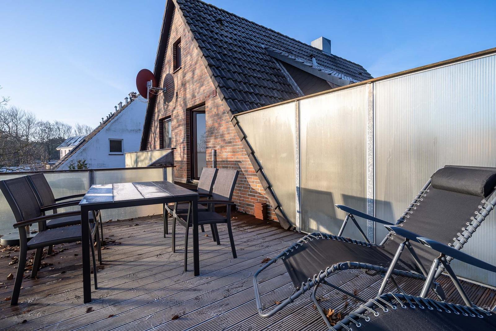 Terrasse mit Tisch, Stühlen und Liegen. Hintergrund: Backsteinhaus mit Dach und Satellitenschüssel.