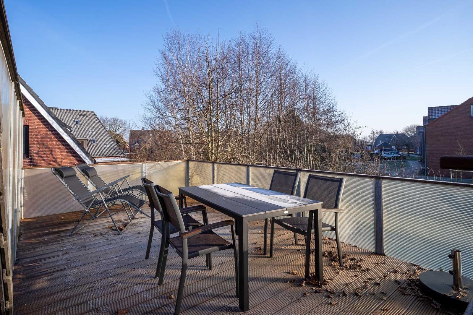 Terrasse mit Tisch, Stühlen und Liegestühlen. Blick auf Bäume und Häuser.