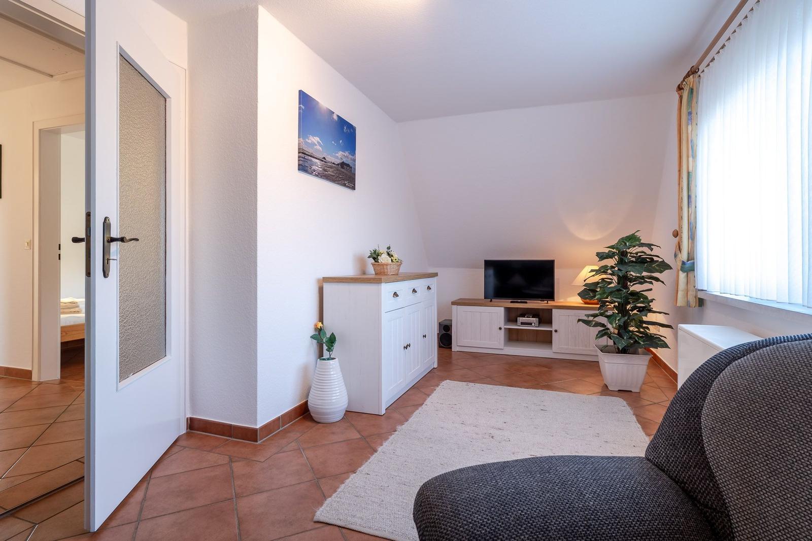 Wohnzimmer mit TV, Pflanzen und Tür zum Schlafzimmer. Terrakottafußboden und weiß lackierte Möbel.