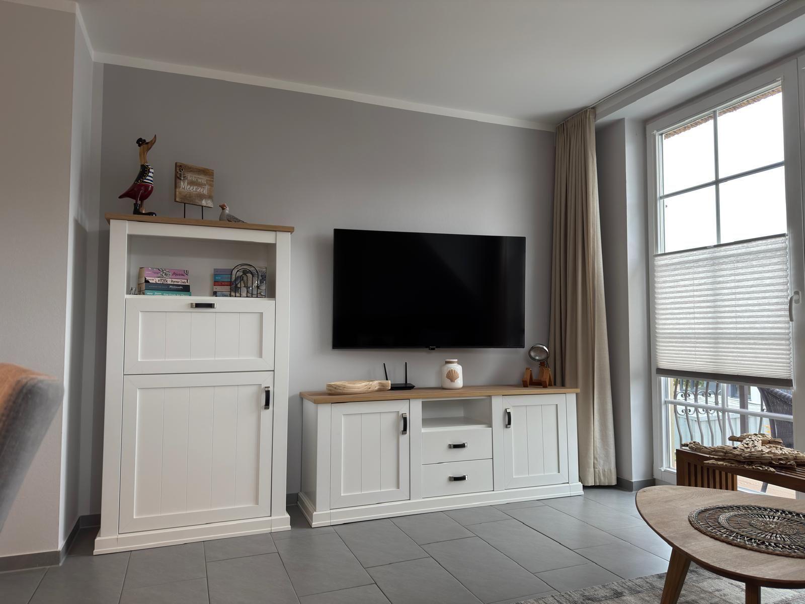 Wohnzimmer mit Fernseher, weißen Schränken und großem Fenster.