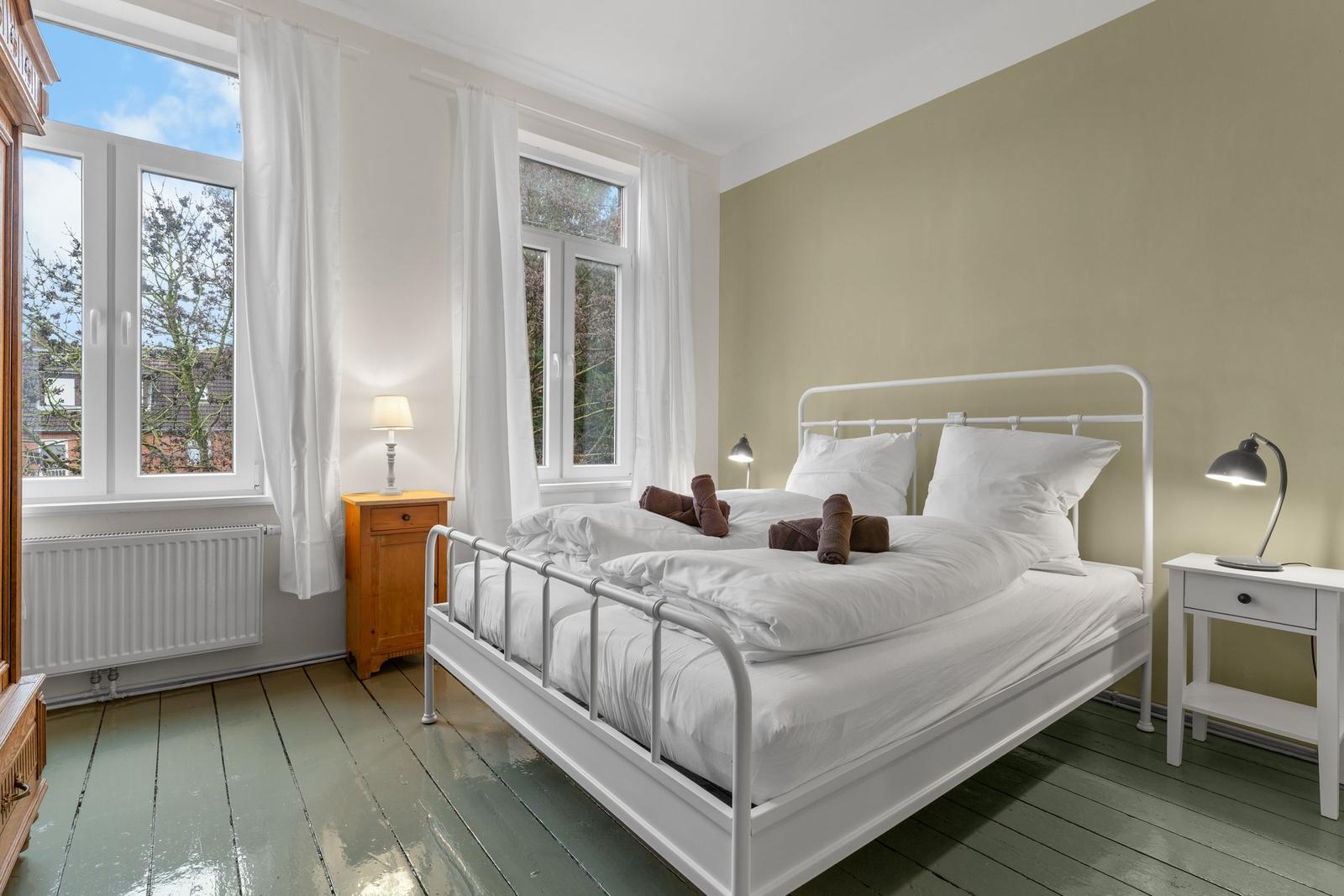 Schlafzimmer mit weißem Bett, zwei Nachttischen und großen Fenstern.