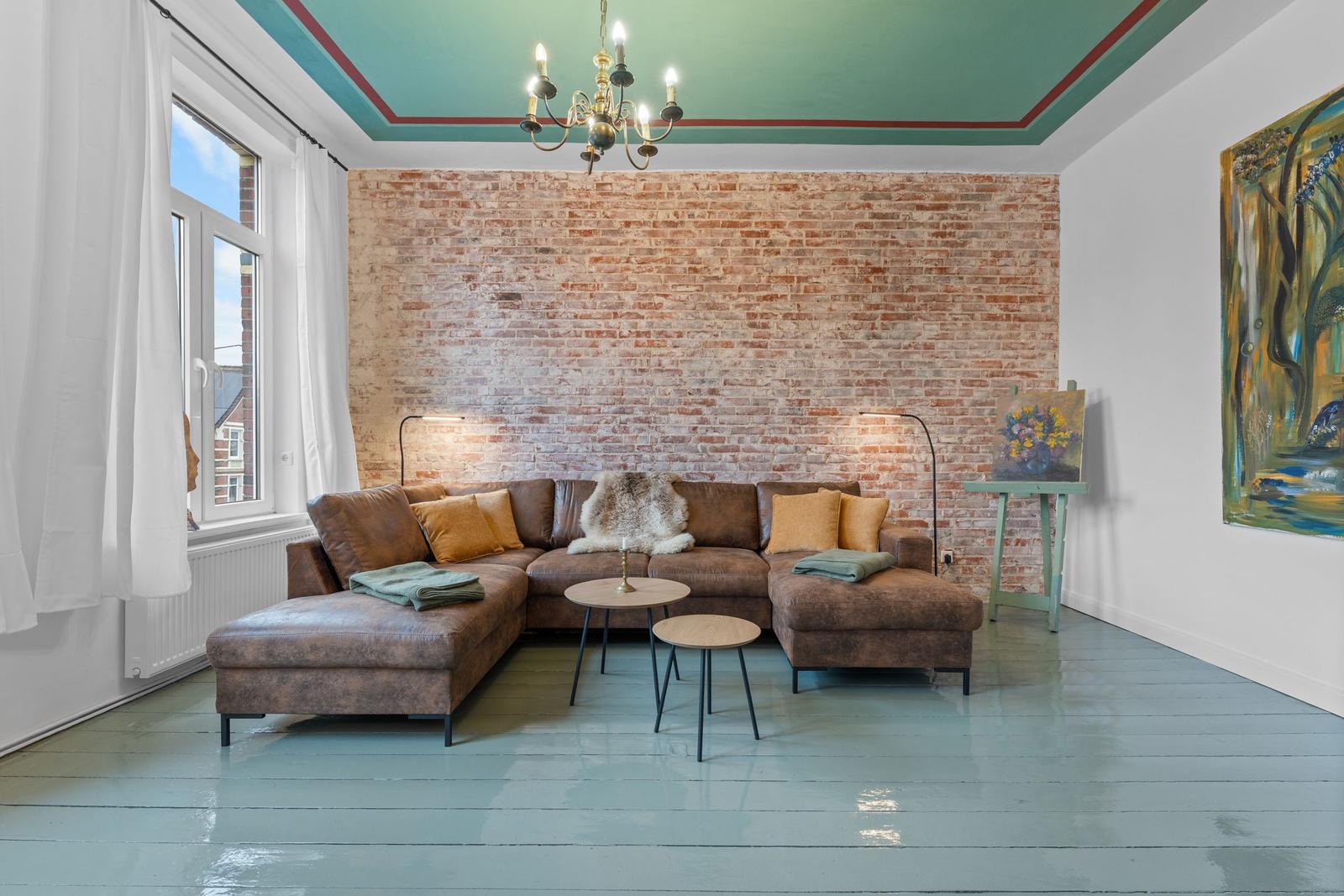 Ein Wohnzimmer mit braunem Sofa, Backsteinwand und grünem Boden. Decke mit Kronleuchter.