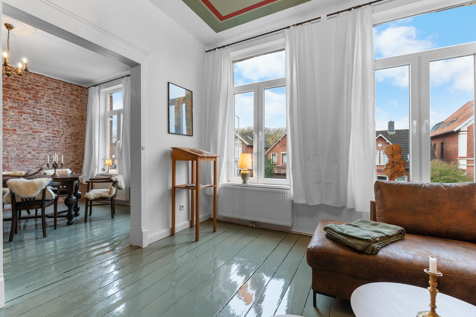 Wohnzimmer mit braunem Sofa und Blick auf die Straße. Holzfußboden und Fenster mit weißen Vorhängen.