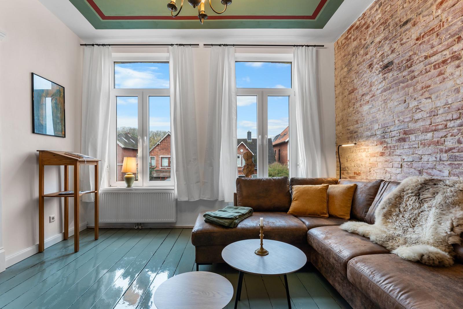 Wohnzimmer mit braunem Sofa, Holzfußboden und Backsteinwand. Große Fenster mit Blick auf Dächer.