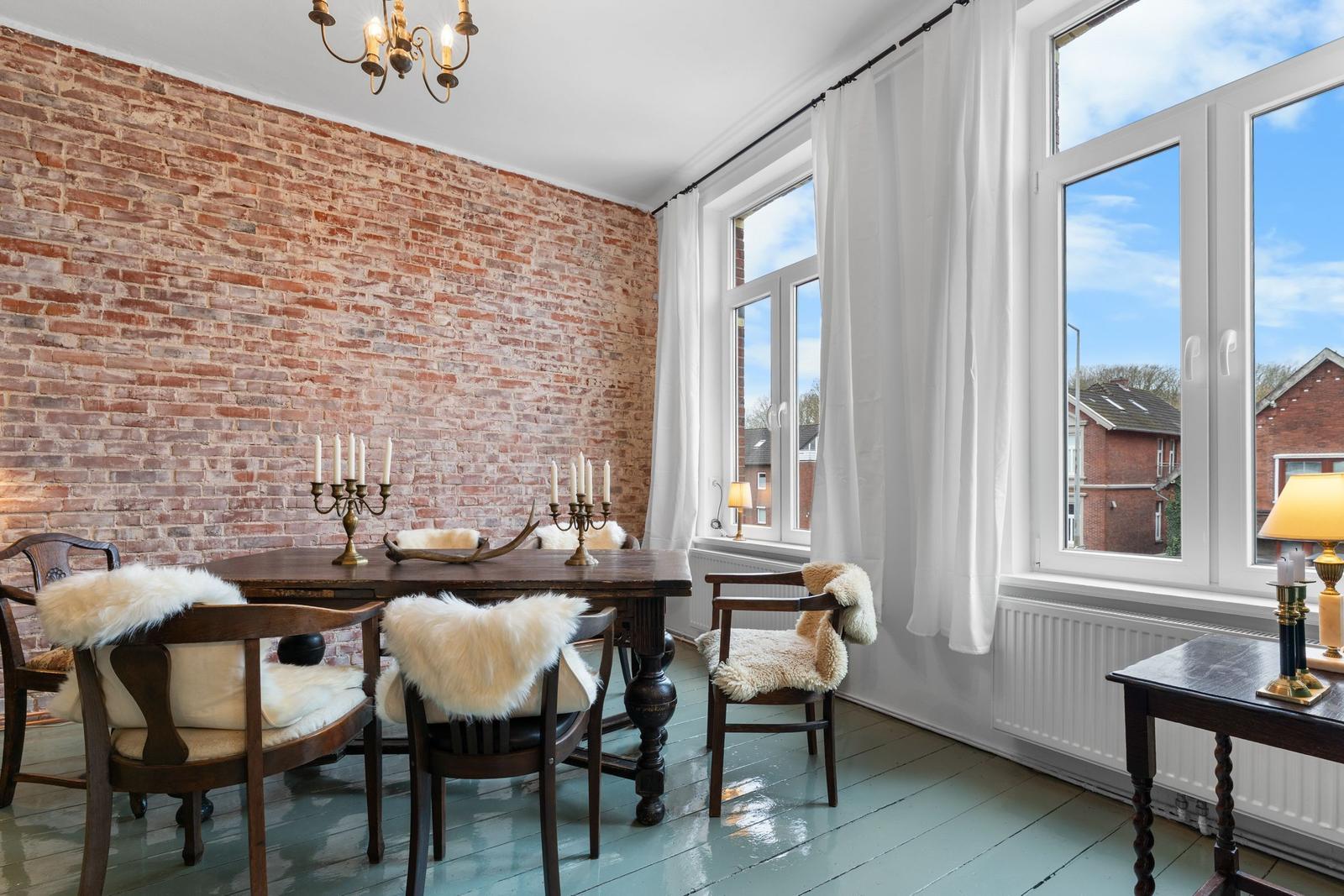 Esszimmer mit rustikalem Holztisch, Stühlen mit Fellkissen und Backsteinwand. Große Fenster mit weißen Vorhängen.