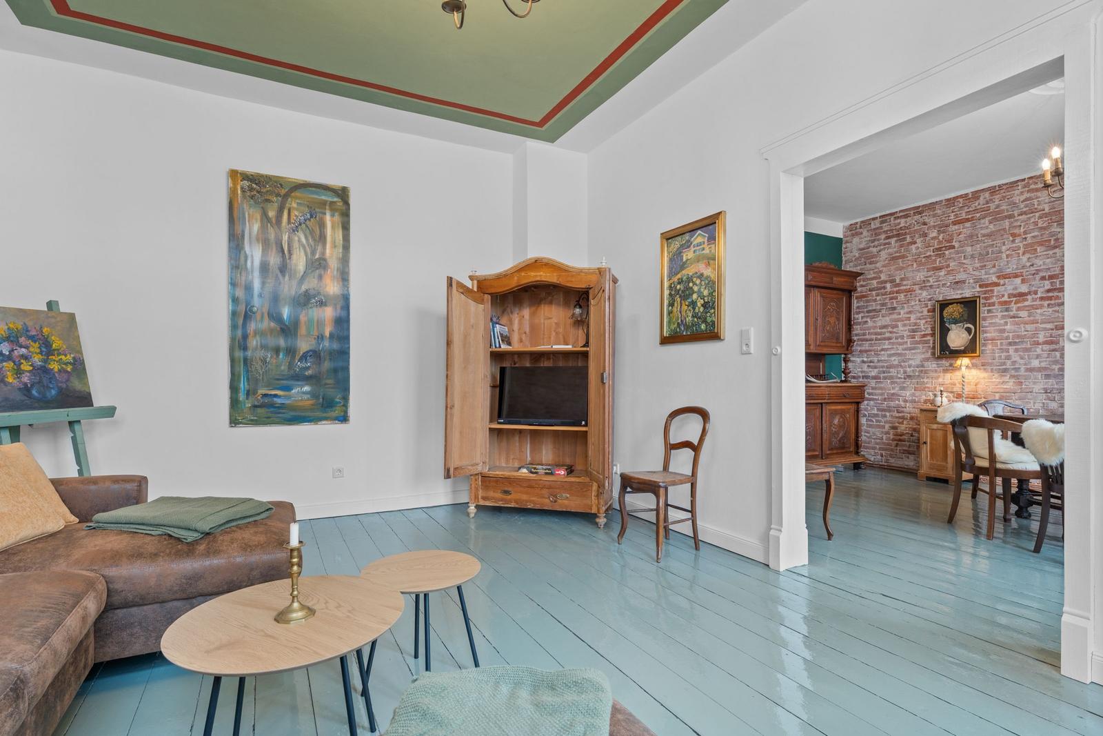 Wohnzimmer mit Holzschrank, Sofa und Gemälden. Durchgang zu Esszimmer mit Backsteinwand.