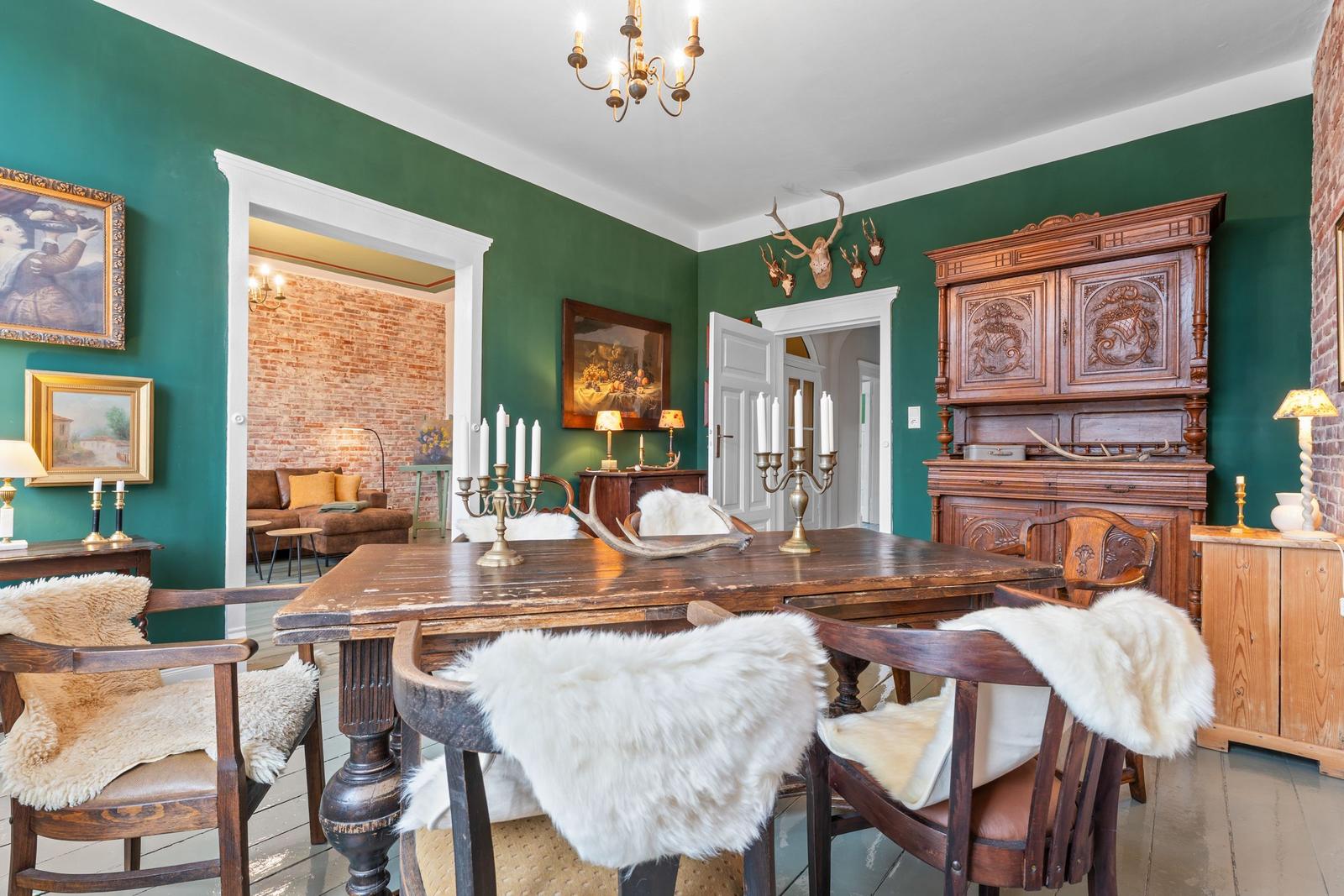 Esszimmer mit Holztisch, Stühlen mit Fell und antikem Schrank an grüner Wand.