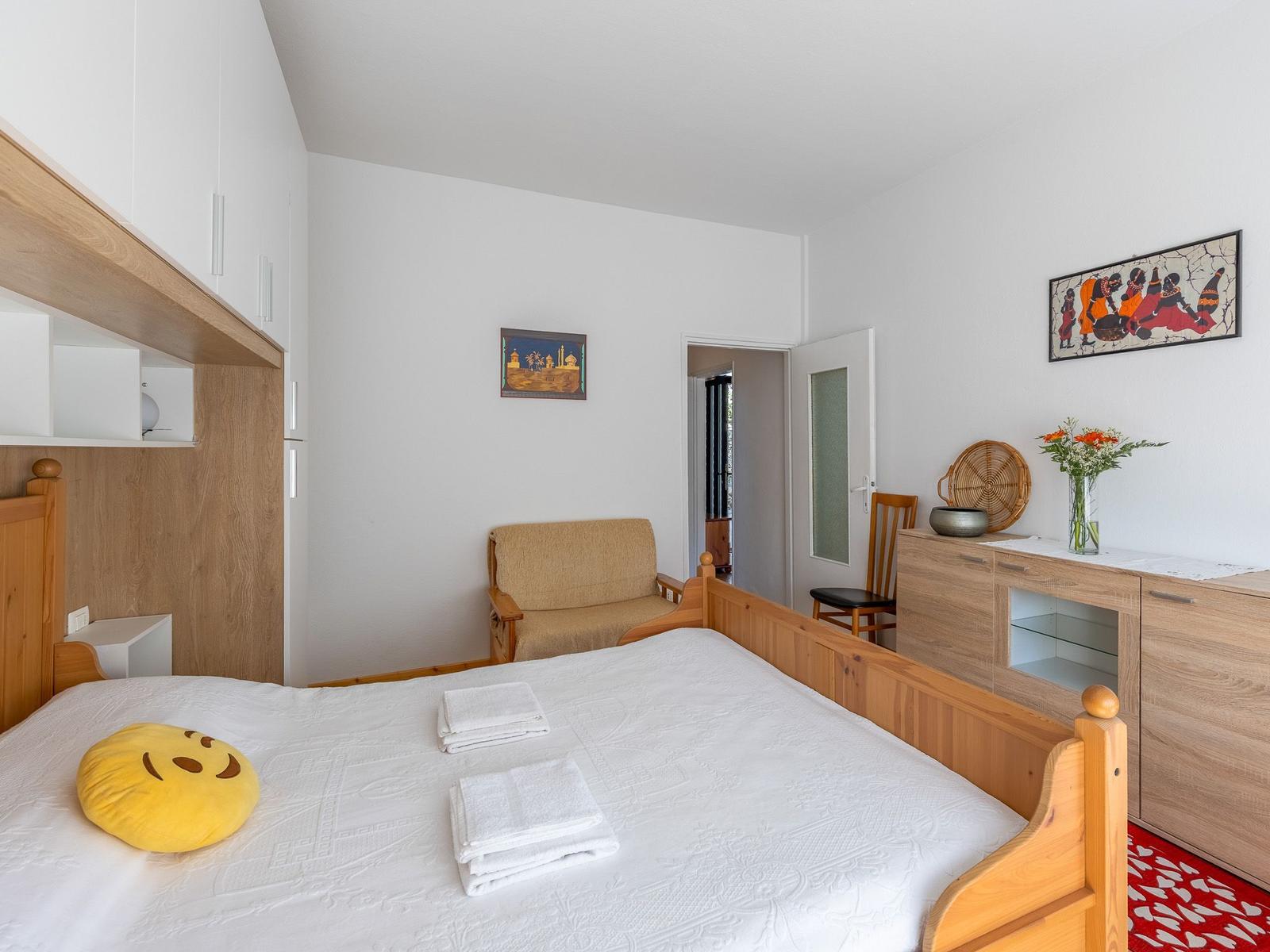 Schlafzimmer mit Holzbett, Kleiderschrank und Stuhl. Auf Bett liegen Handtücher und Kissen.