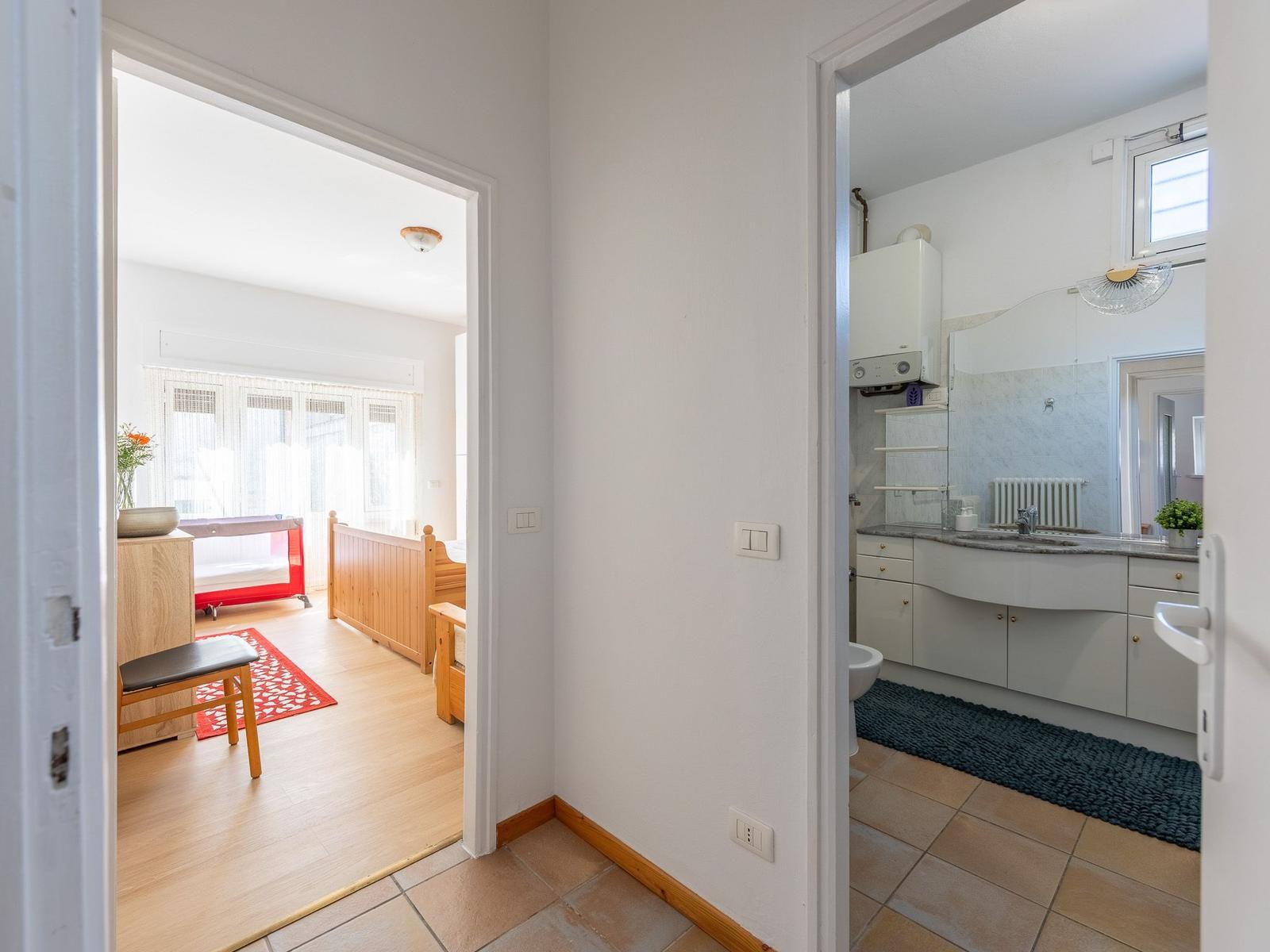 Zimmer mit Holzboden und Babybett. Nebenraum ist Bad mit Waschbecken und Spiegel.