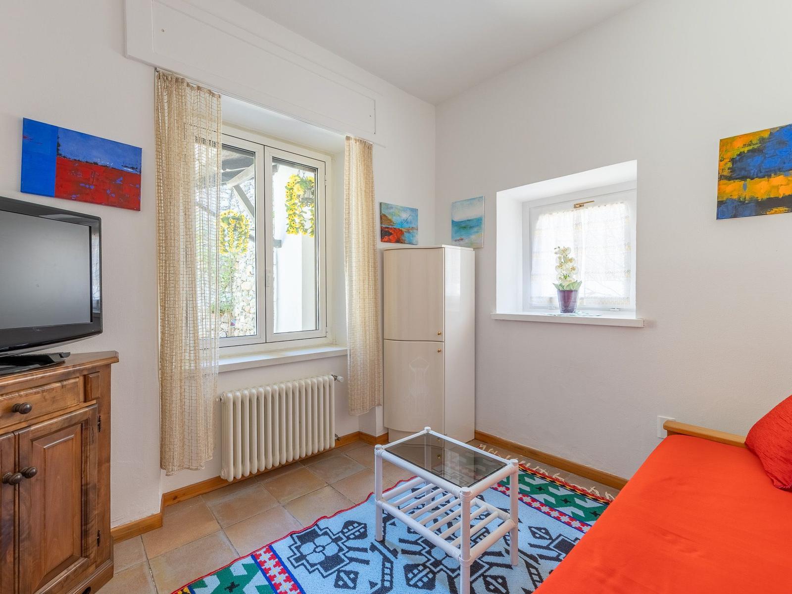 Zimmer mit Fenster, Schrank, TV und Bett mit roter Bettwäsche.