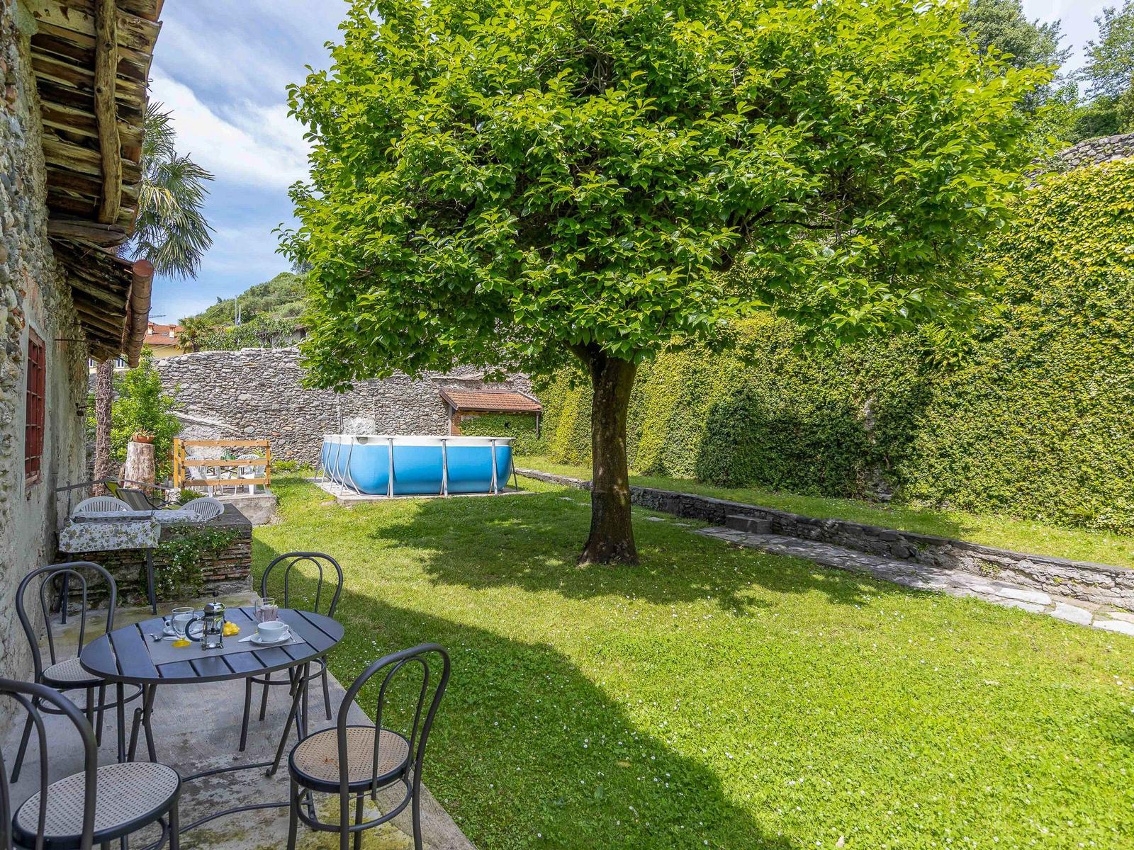 Hinterhof mit grünem Rasen, Baum, Pool und Terrasse mit Tisch und Stühlen.