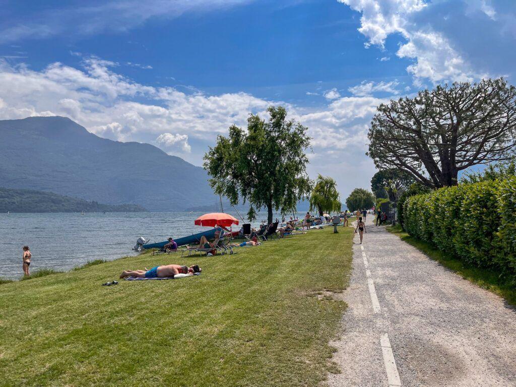 Grüner Strandabschnitt am See mit Sonnenschirmen und Bäumen. Menschen entspannen sich am Ufer.