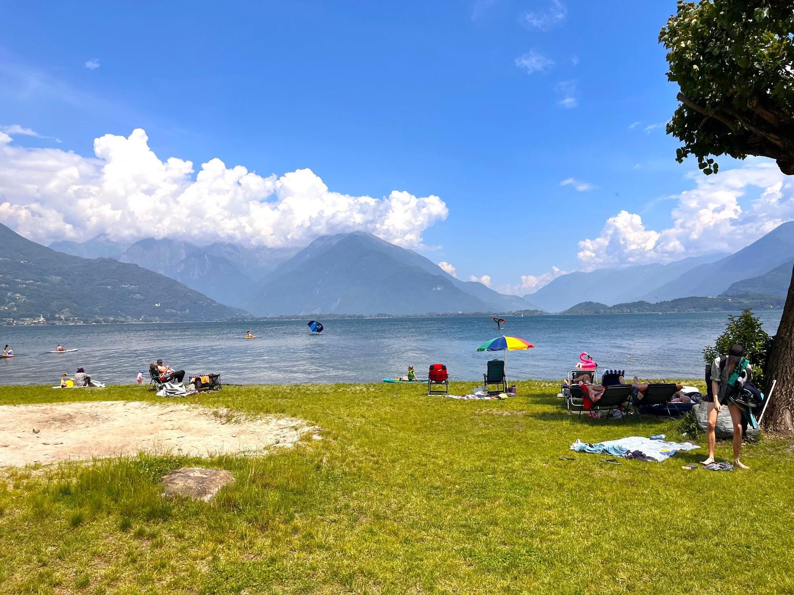 Grüner Strand mit Liegestühlen und buntem Sonnenschirm am See. Im Hintergrund sind Berge und Wassersportler zu sehen.