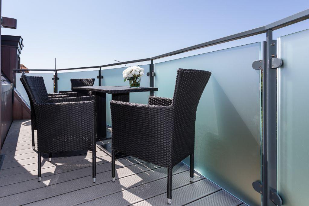 Terrasse mit Tisch und vier Stühlen, Glasgeländer und Blumenarrangement.