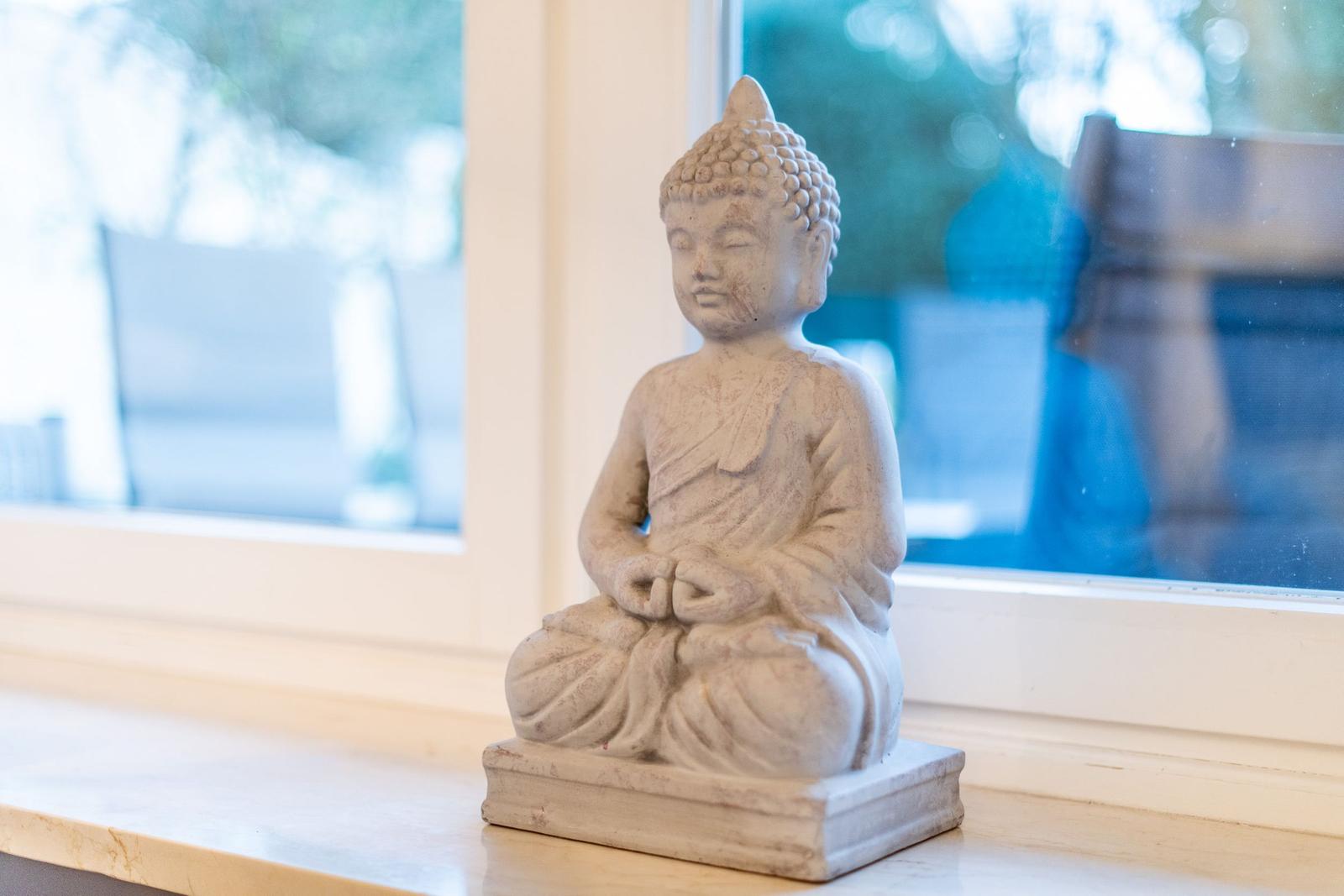 Eine Buddha-Statue sitzt auf einem Fensterbrett. Im Hintergrund ist ein Fenster mit Blick nach draußen.