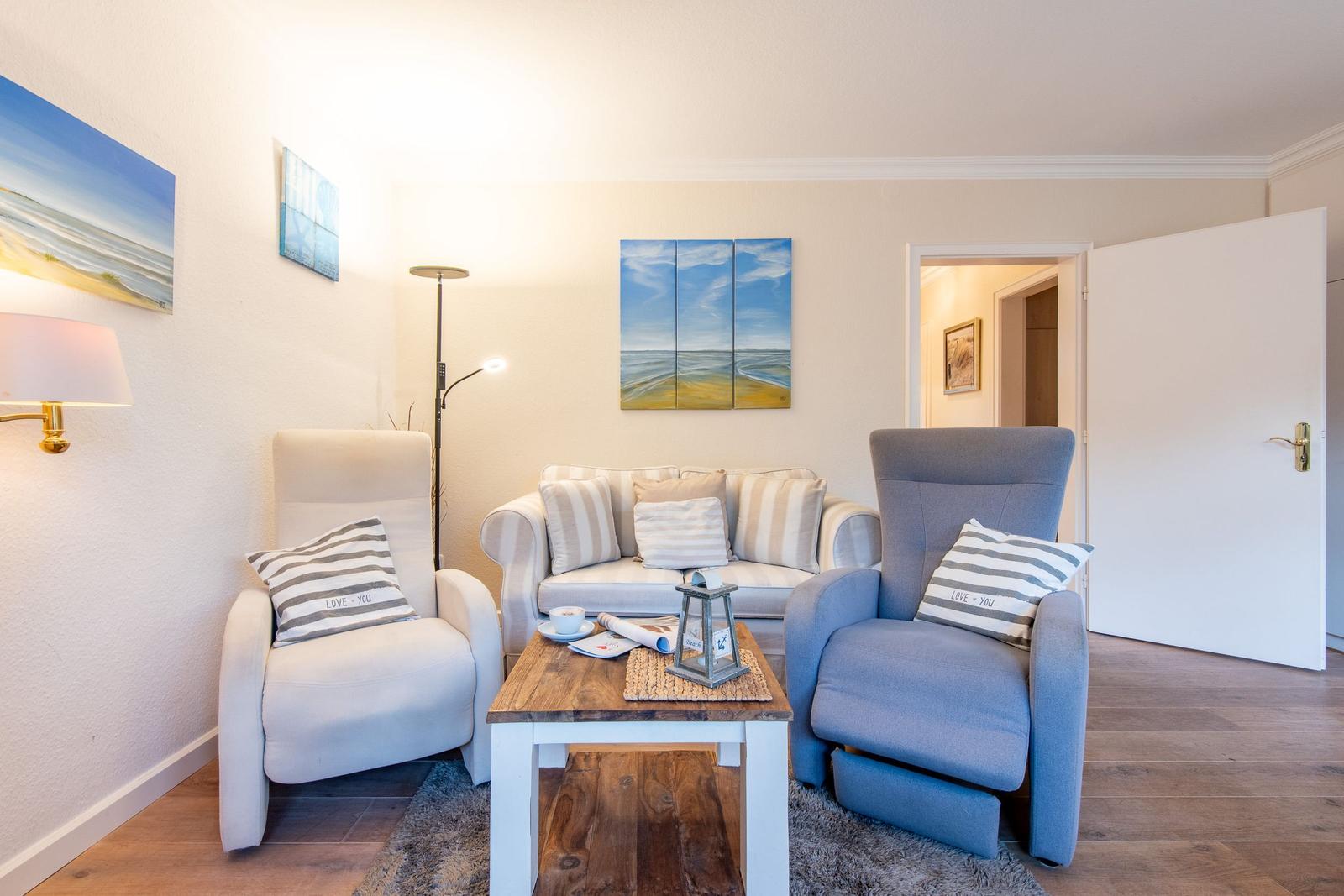 Wohnzimmer mit Sofa, zwei Sesseln und Holztisch. Wandbilder zeigen Strandlandschaften.