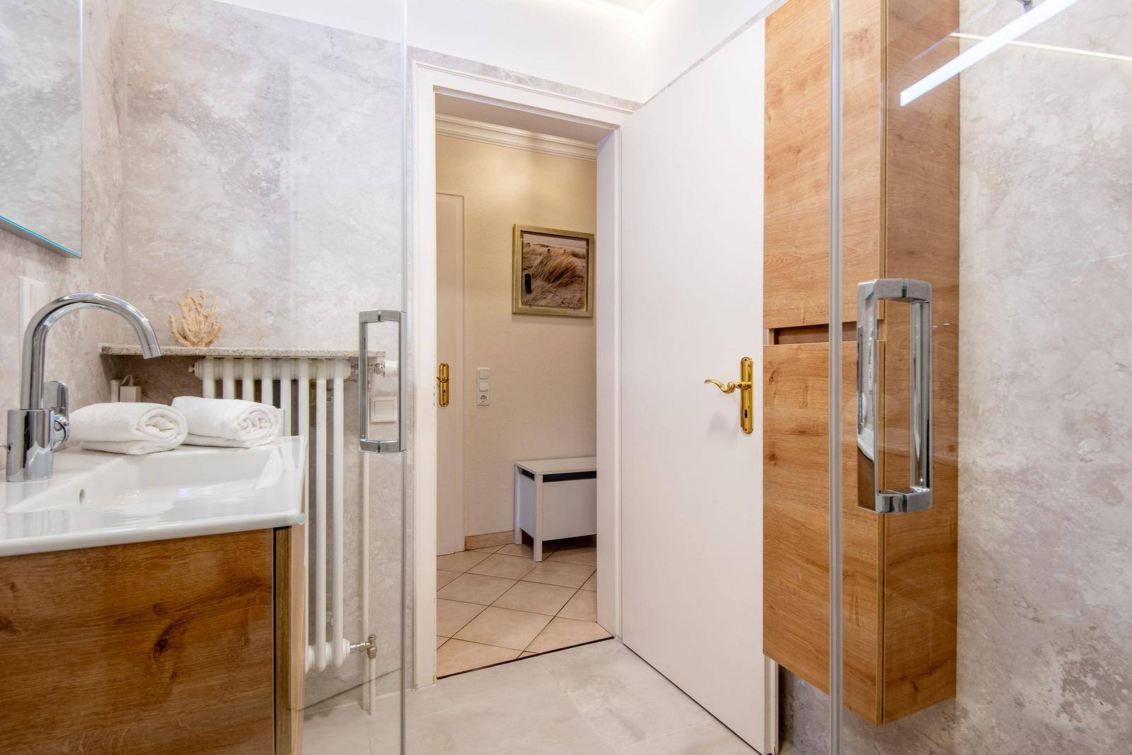 Badezimmer mit Dusche, Waschbecken und Holzregalen. Tür führt in Wohnbereich.