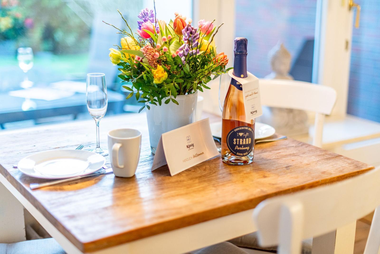 Tisch mit Blumen, Flasche und Geschirr vor Fenster.