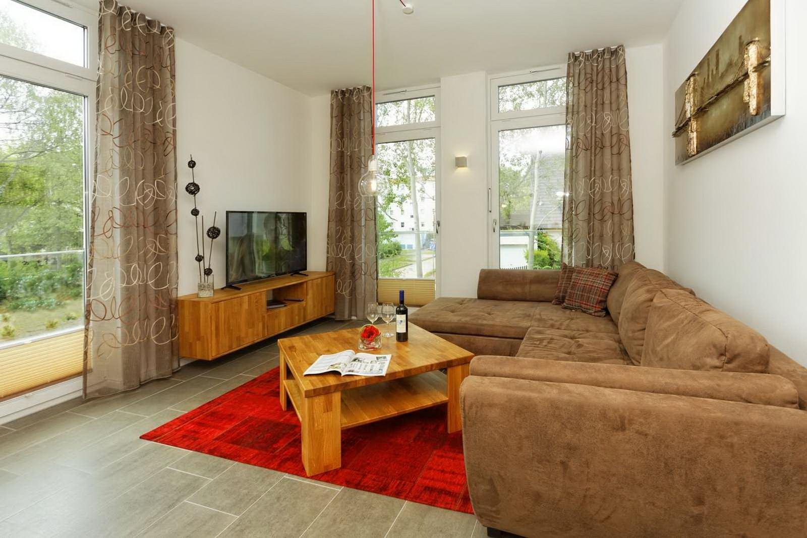 Wohnzimmer mit braunem Sofa, Holztisch und Fernseher. Große Fenster mit Blick auf Garten.