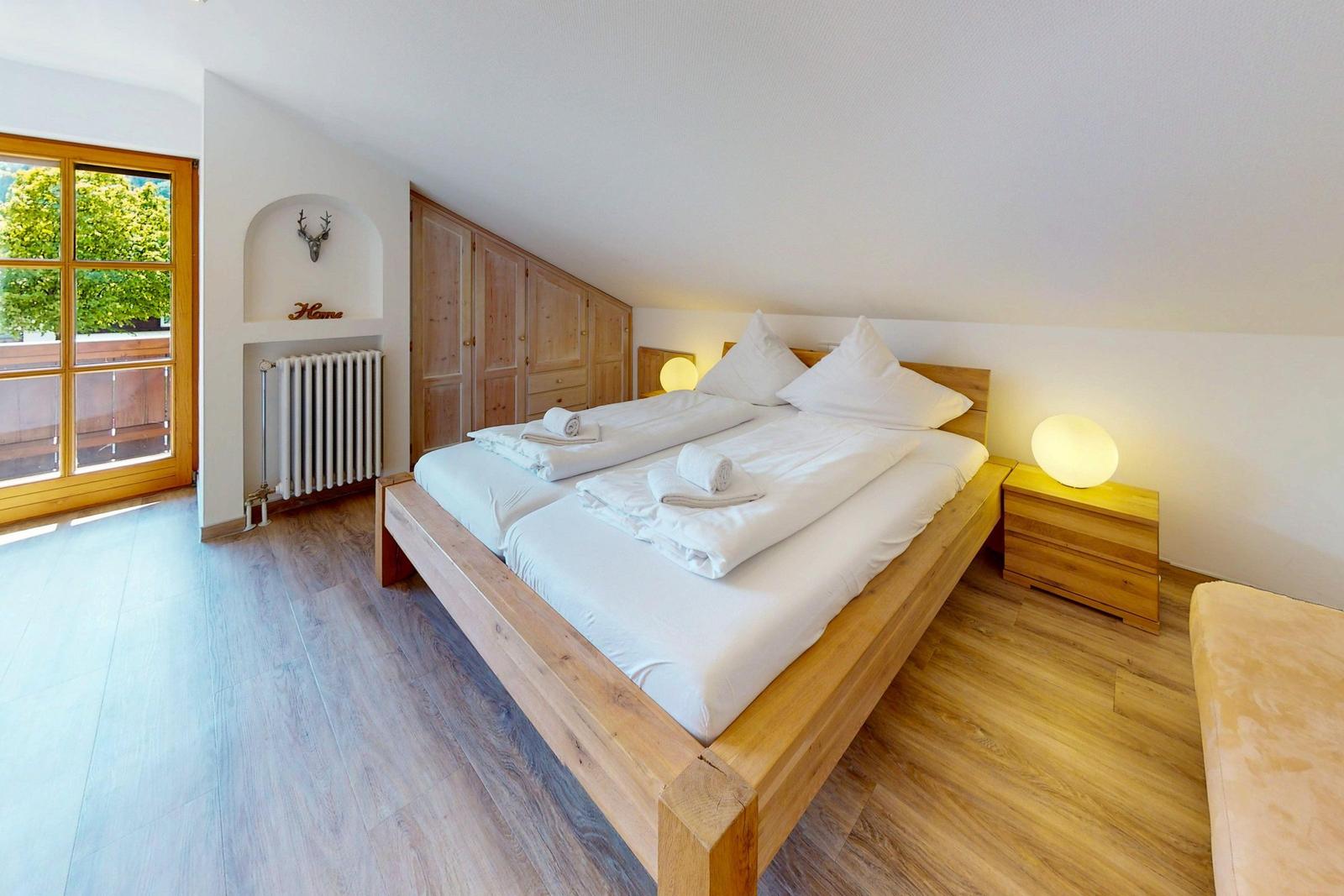 Schlafzimmer mit Holzbett, Nachttisch und Fenster. Holzschrank und Heizkörper sind sichtbar.