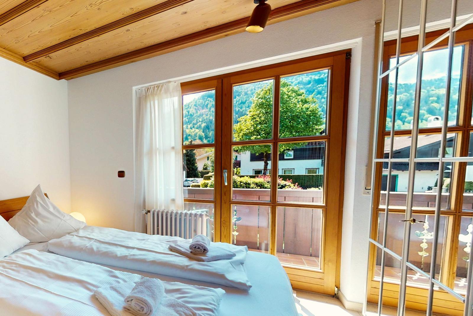 Schlafzimmer mit Blick auf grüne Landschaft und Balkon. Holzdecke und weißes Bett.