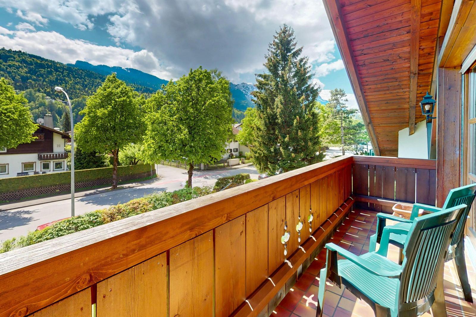Terrasse mit Holzgeländer und zwei grünen Stühlen. Blick auf Straße, Bäume und Berge.