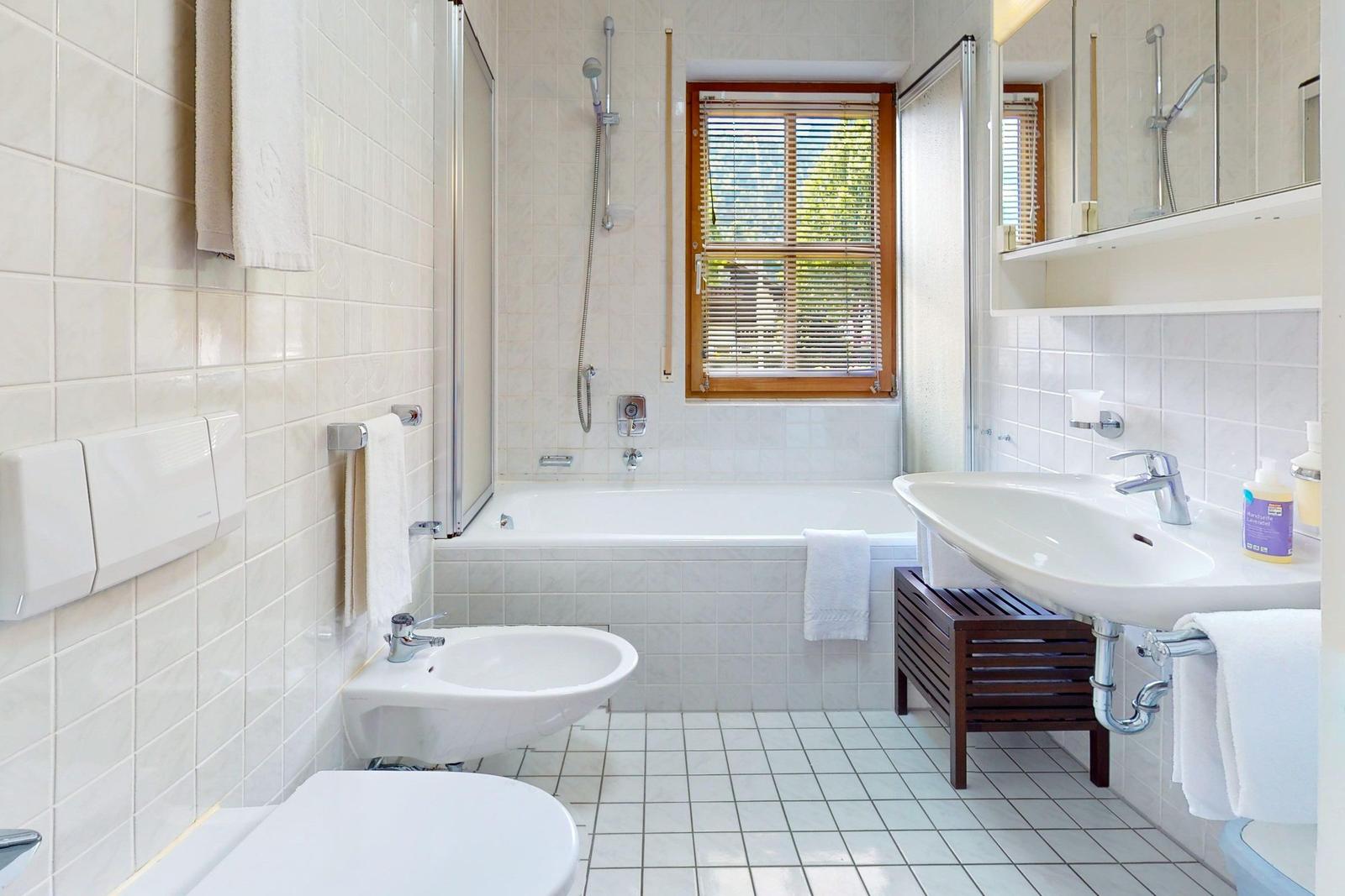 Badezimmer mit Badewanne, Dusche, Waschbecken und Toilette. Holzfenster mit Jalousie.