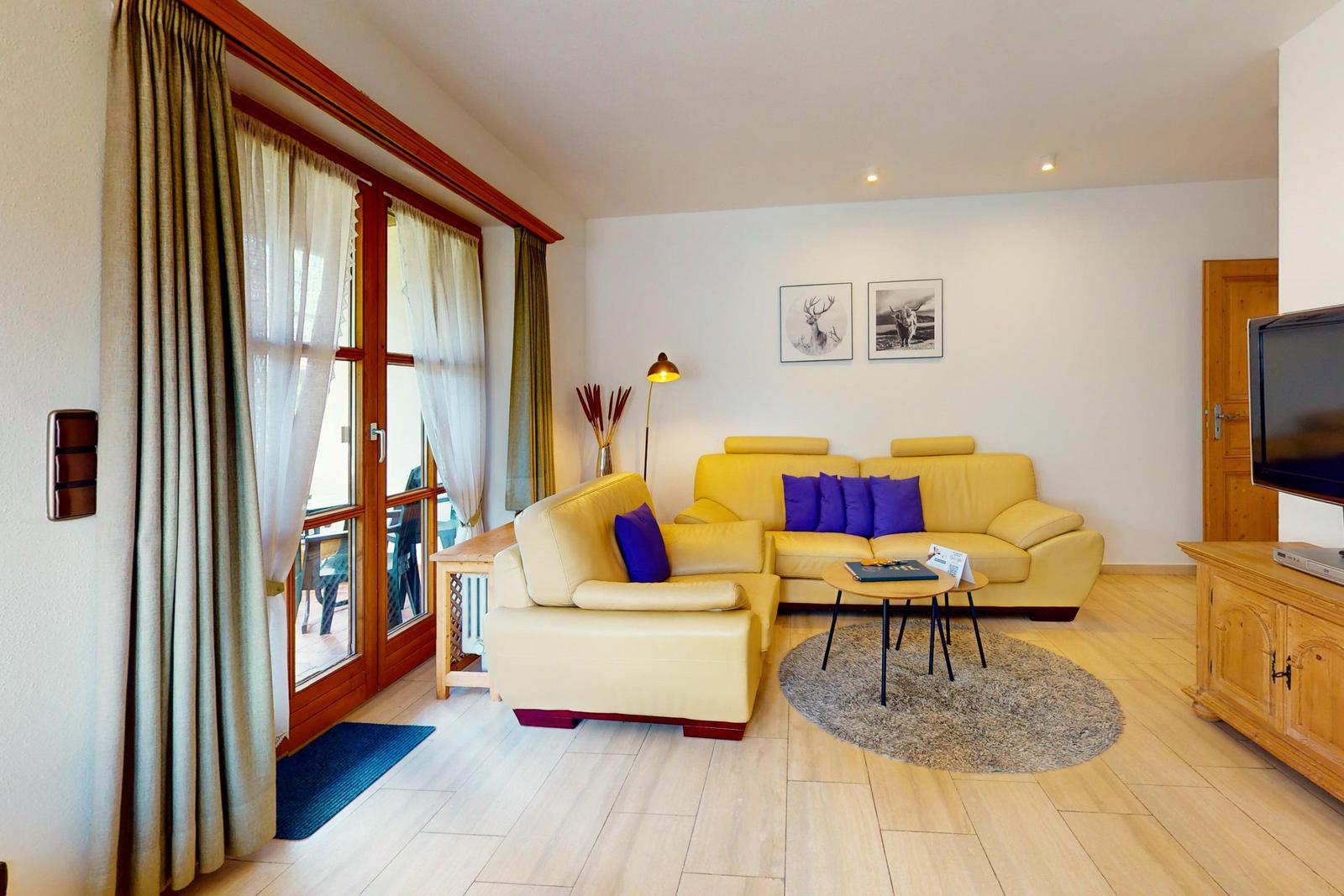 Wohnzimmer mit gelbem Sofa, runden Teppich und Holzboden. Fenster mit Vorhängen und TV-Möbel.