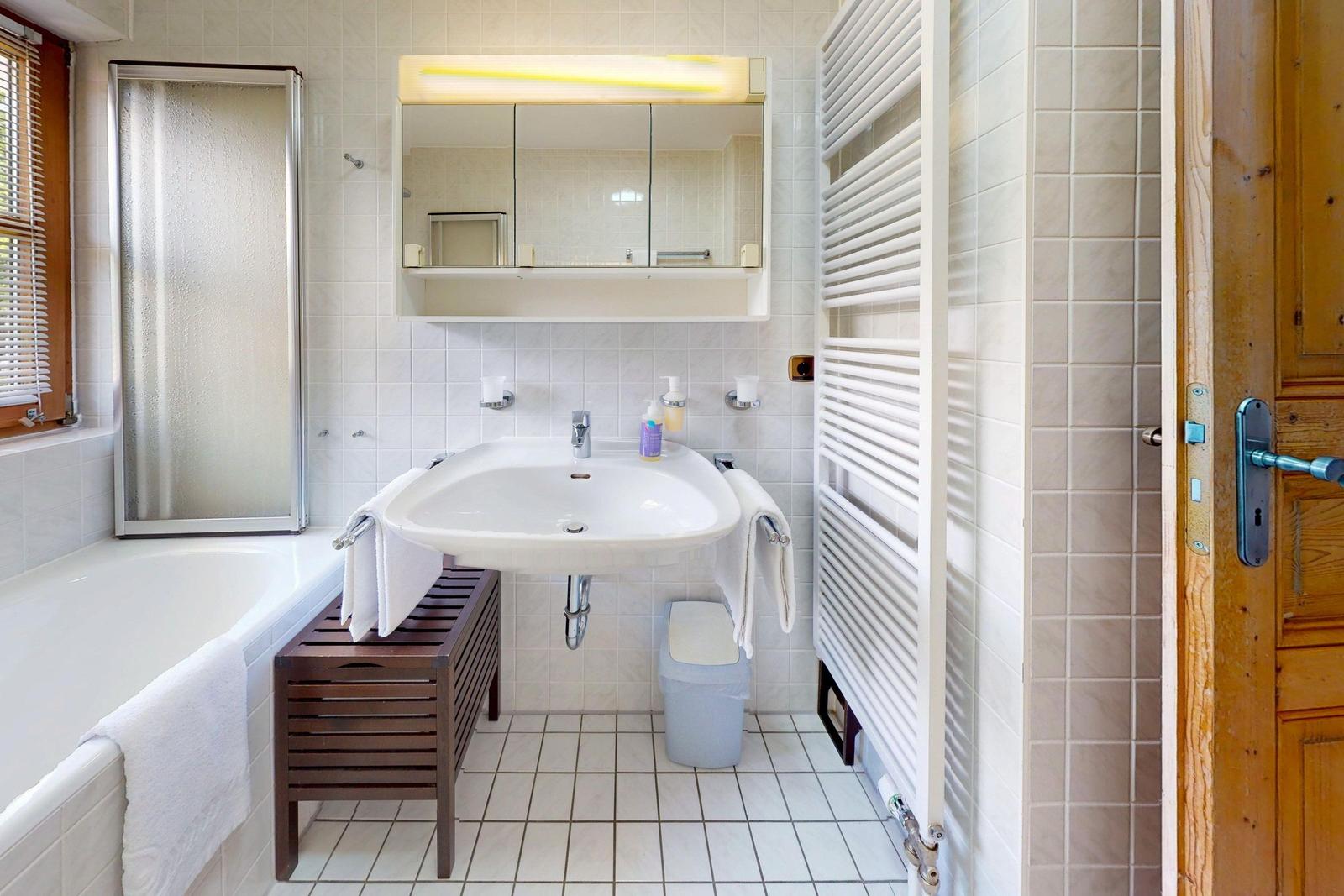 Badezimmer mit Badewanne, Waschbecken und Spiegel. Heizkörper und Holztür sind sichtbar.
