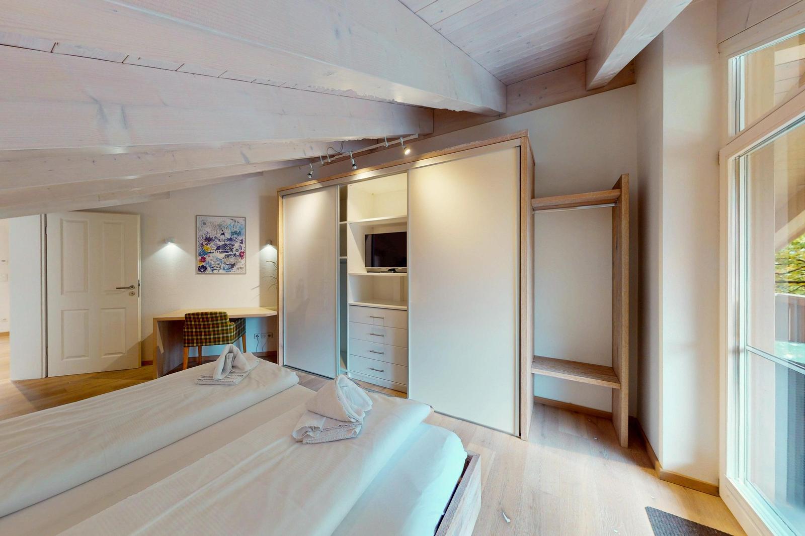 Schlafzimmer mit Doppelbett, Kleiderschrank und Fenster. Blick auf Balkon und Holzbalkendecke.
