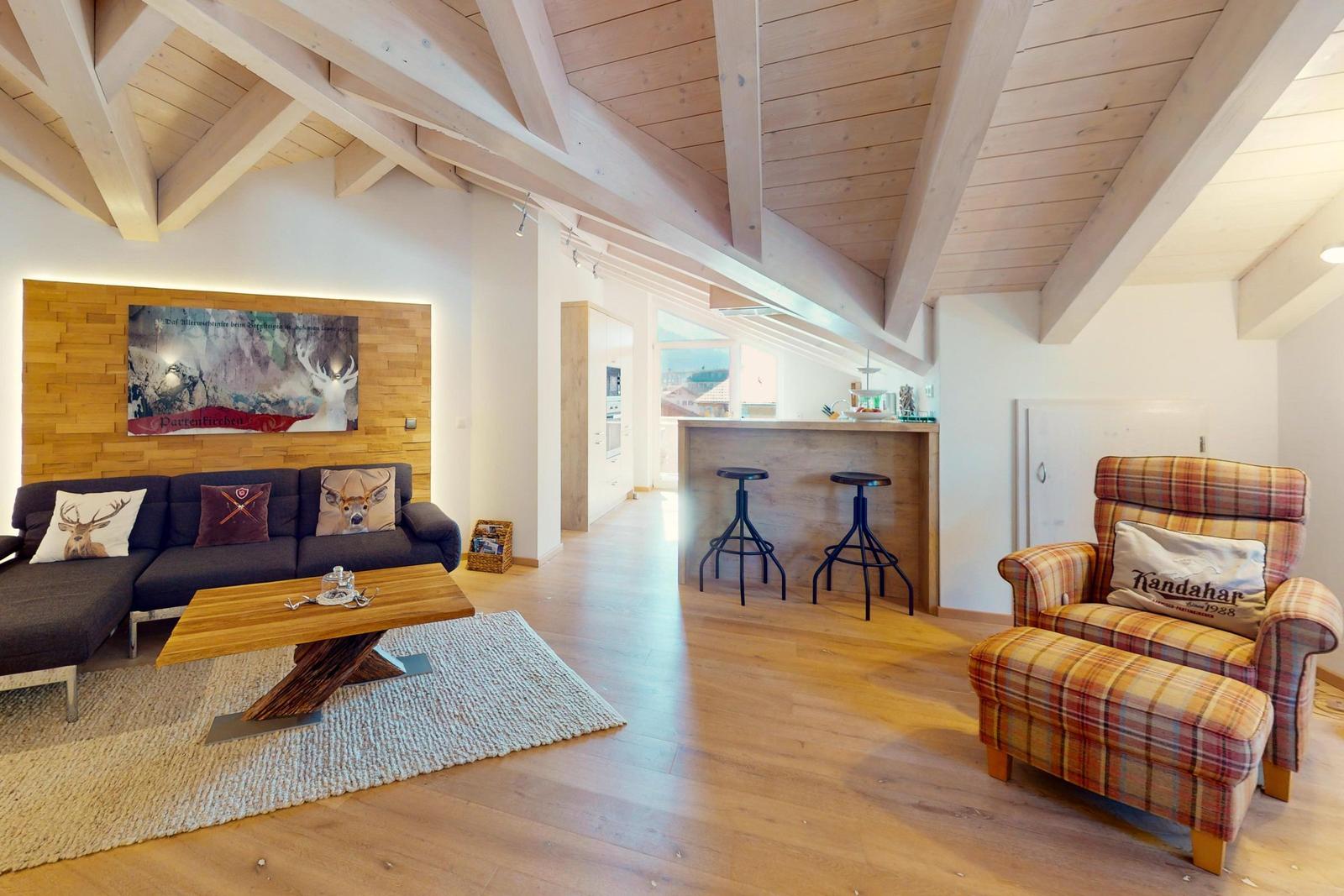 Wohnbereich mit Sofa, Sessel und Küchenbereich unter Holzdecke.