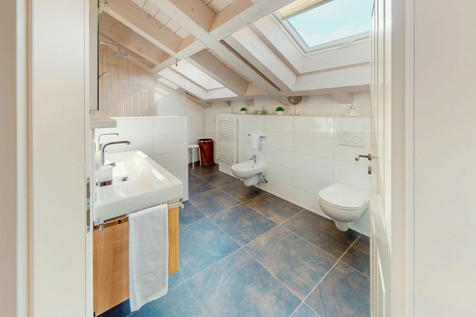 Badezimmer mit Dusche, Toilette und Waschbecken unter Holzdecke mit Dachfenster.