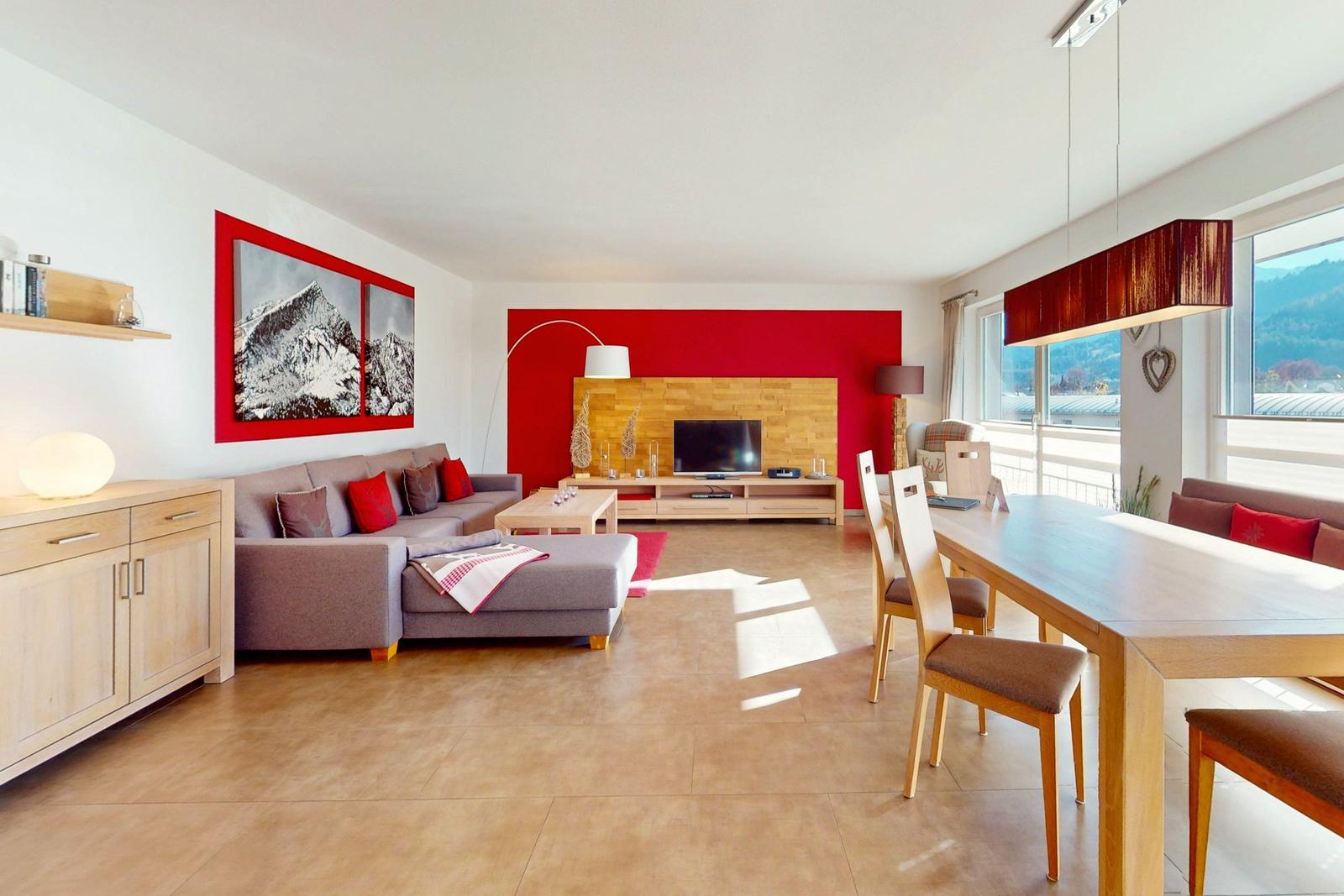 Wohn- und Essbereich mit Sofa, TV und Tisch. Rote Wand und Bergbilder.