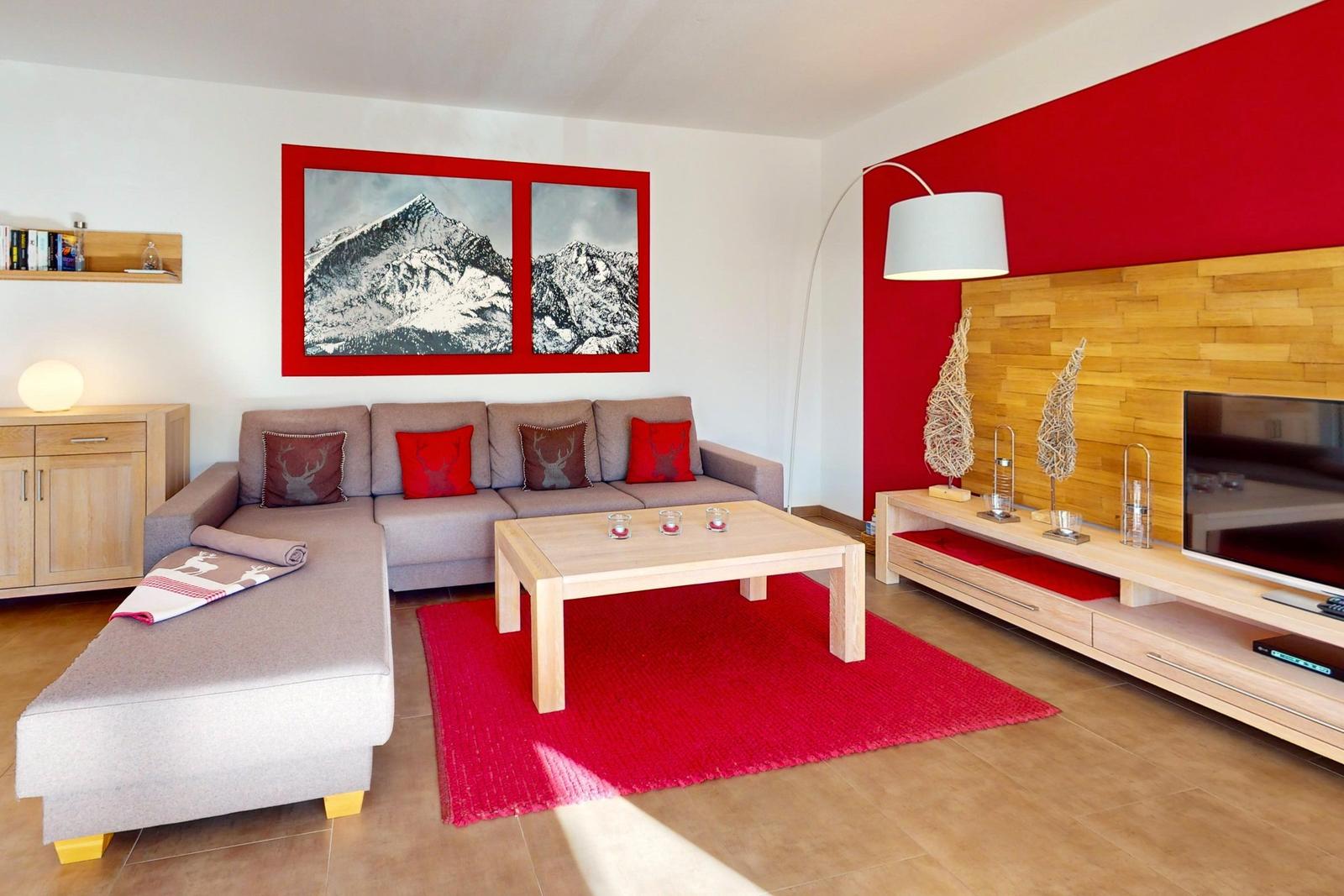 Wohnzimmer mit grauem Sofa, rotem Teppich und Holzmöbeln. Wandbild zeigt Berge.