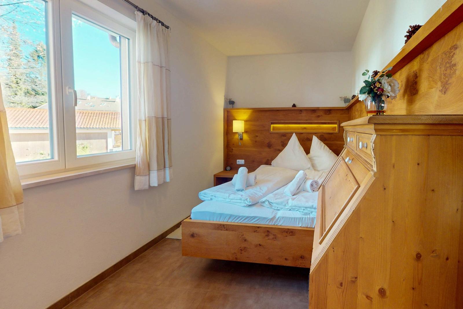 Schlafzimmer mit Holzbett, Fenster und Schrank. Blick auf Dach und Bäume.