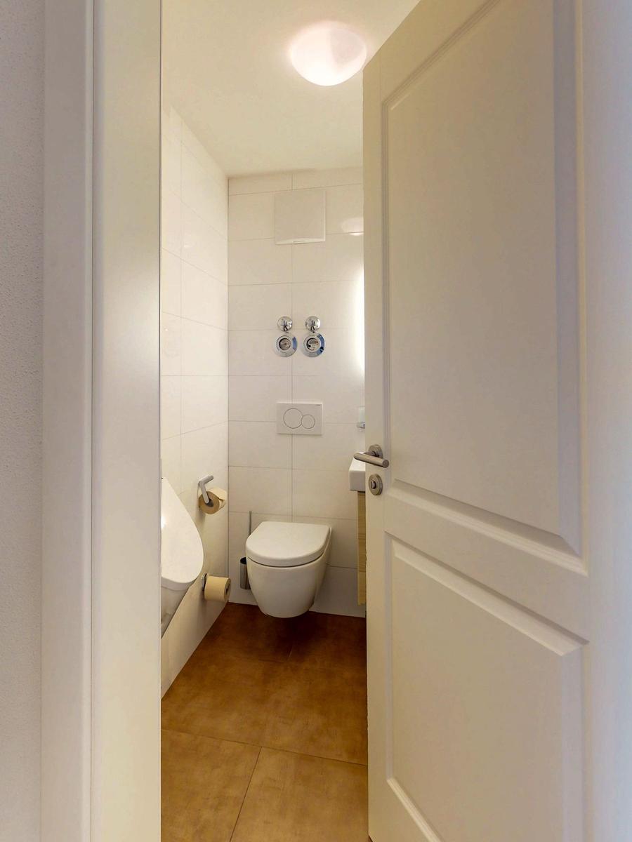 Badezimmer mit Toilette, Dusche und Toilettenpapierhalter. Bodenfliesen und weiße Wände.