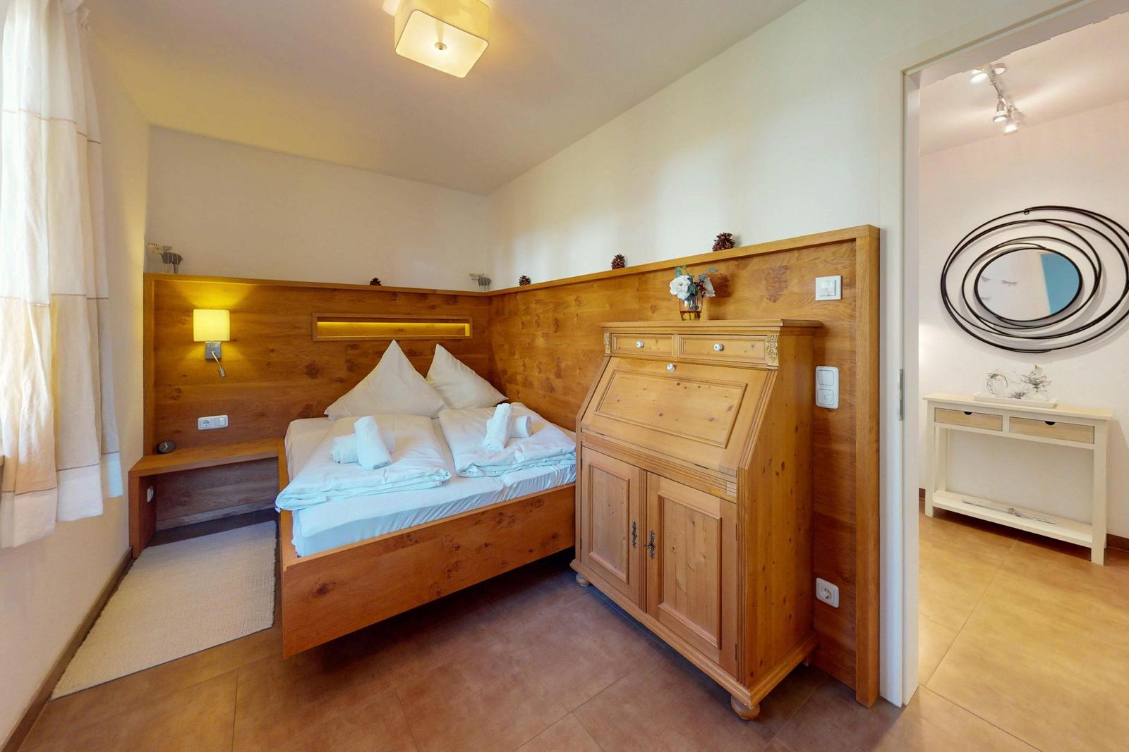 Schlafzimmer mit Holzbett, Schrank und Nachttisch. Tür führt in einen Flur mit Spiegel.