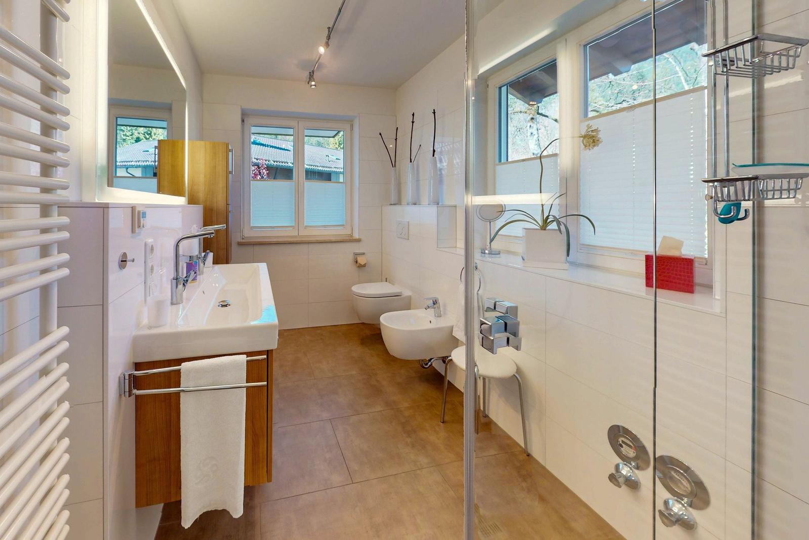 Badezimmer mit Dusche, Waschbecken und Toilette. Fenster mit Jalousie und Pflanze.