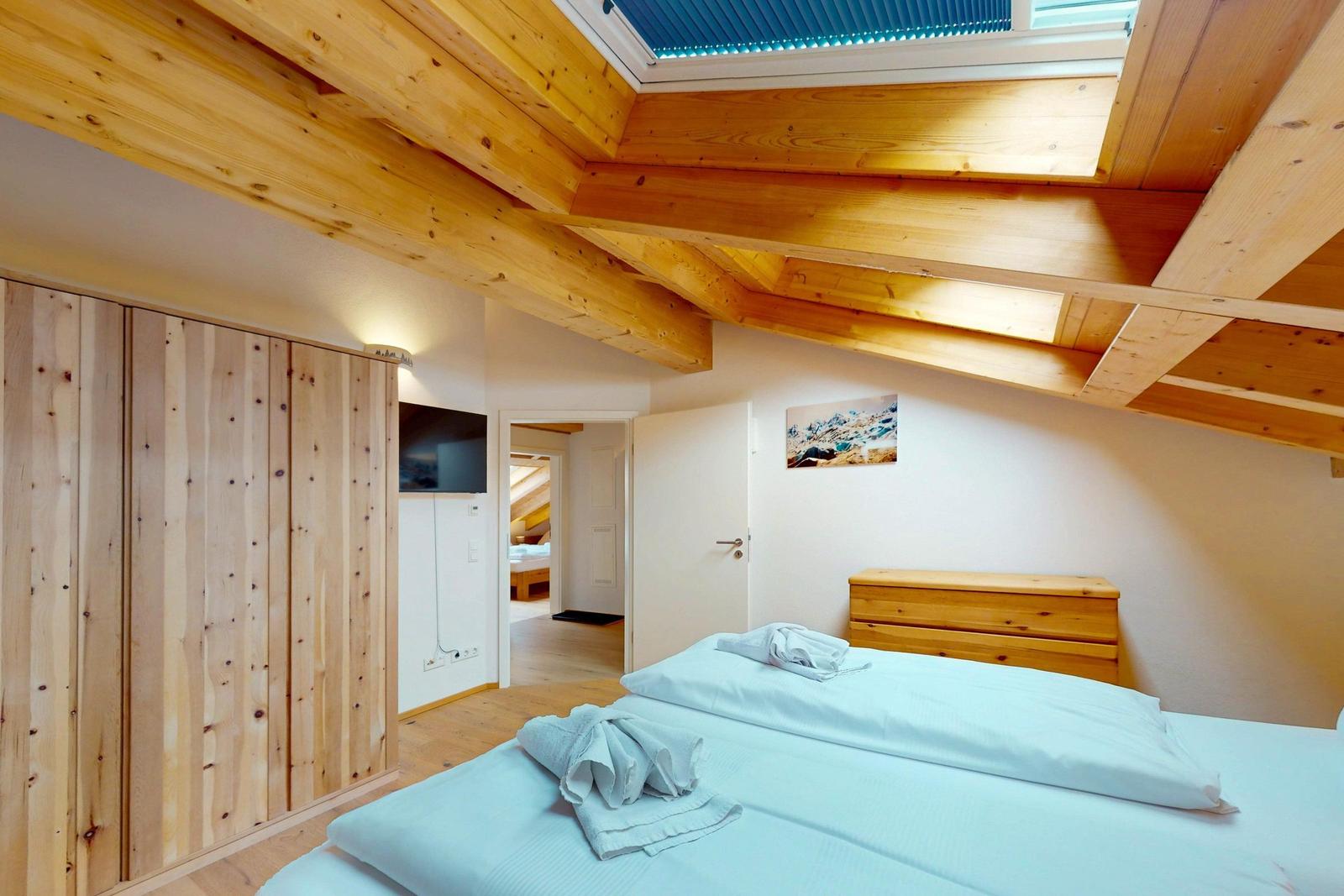Schlafzimmer mit Holzbalkendecke, Bett, Kleiderschrank und TV. Durchgang zu weiteren Räumen.