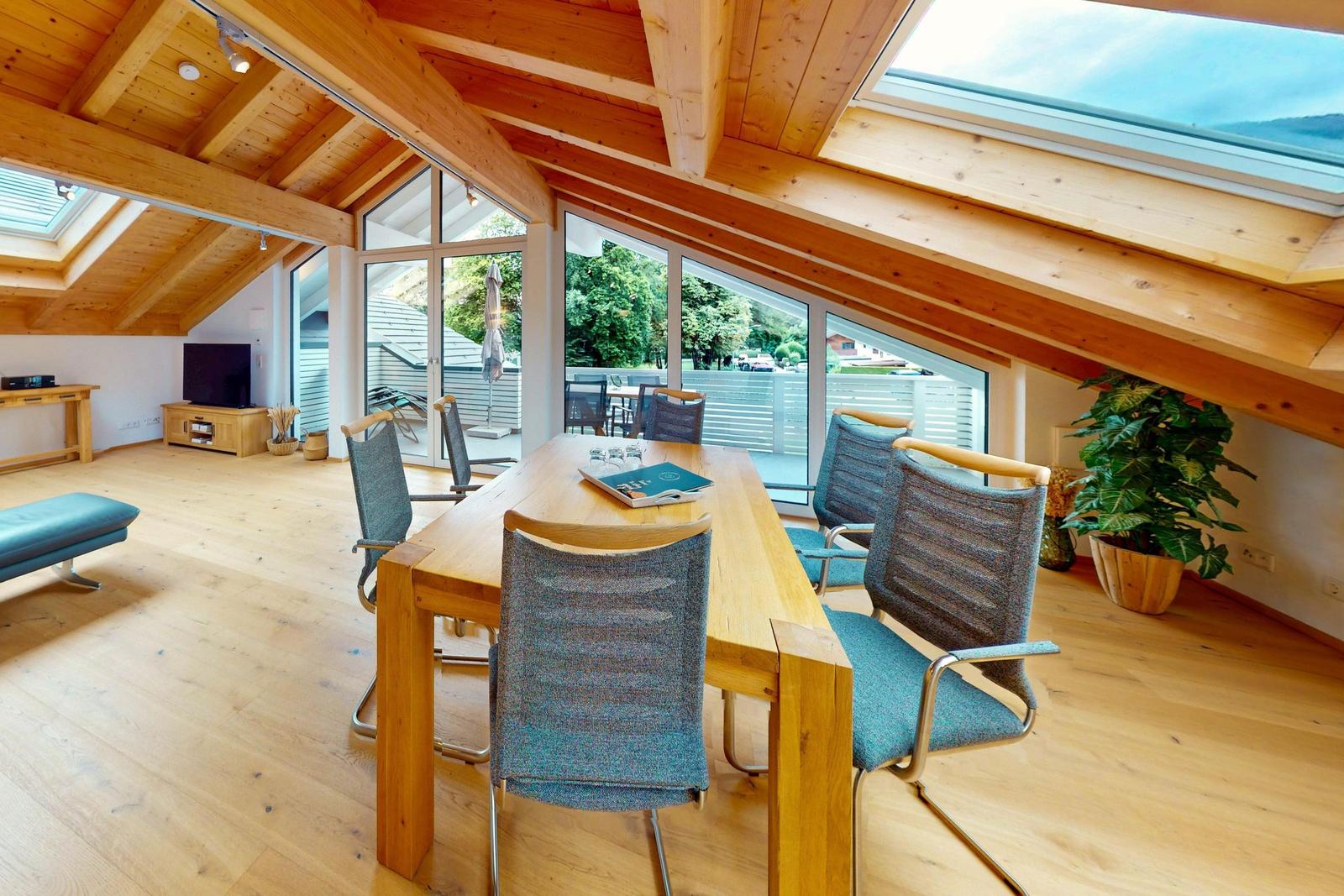 Ein Esszimmer mit Holzdecke, Tisch und Stühlen. Durch große Fenster ist eine Terrasse sichtbar.