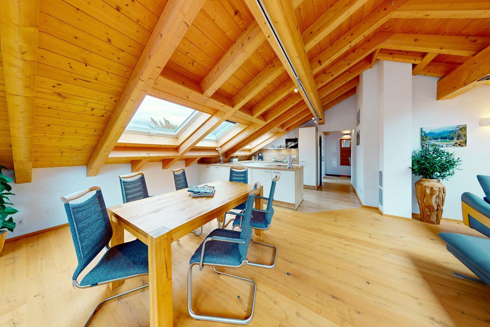 Dining room with wooden table, blue chairs, and kitchen in attic space with wooden beams and skylights.