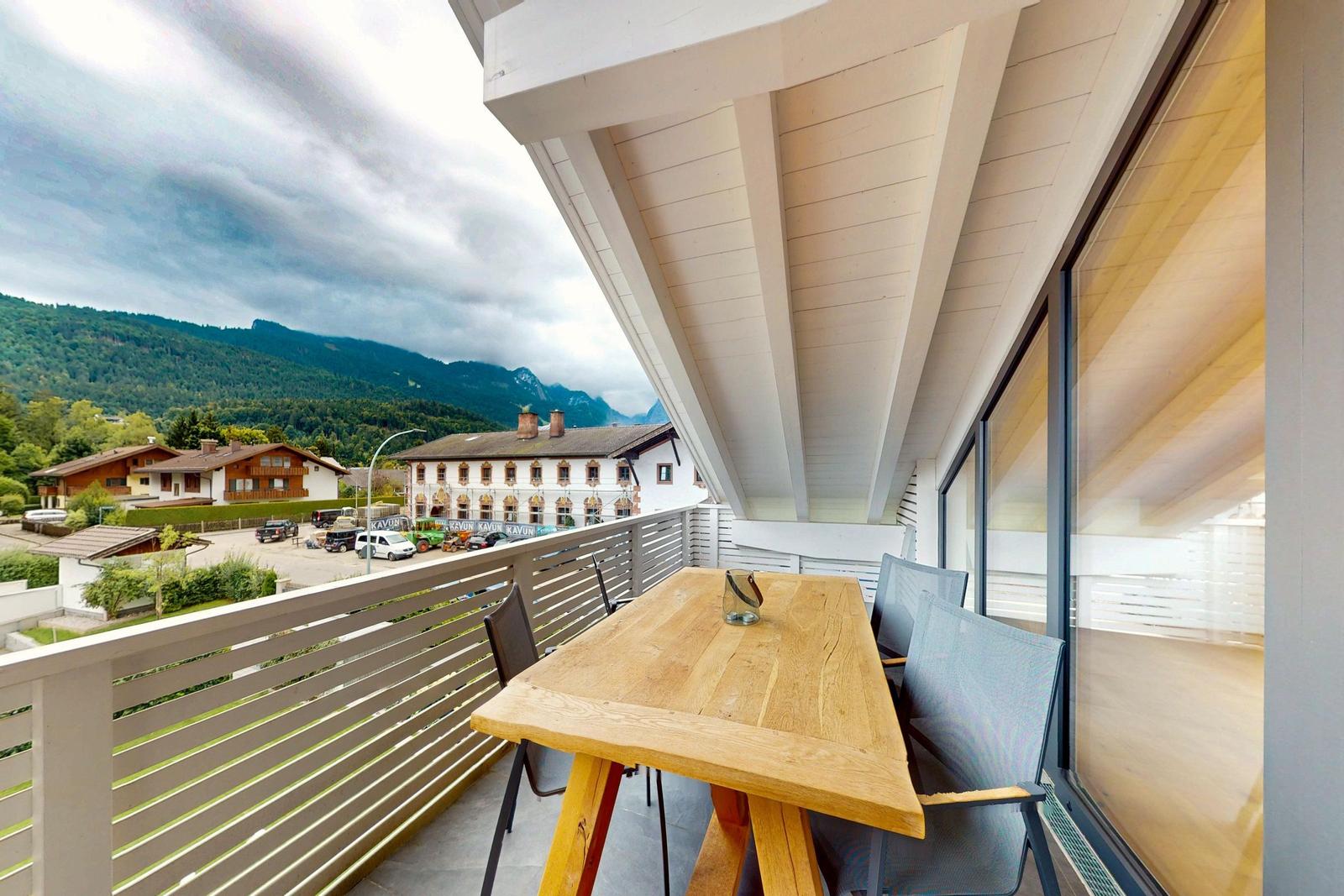 Terrasse mit Holztisch und Stühlen. Blick auf Berge und Gebäude.