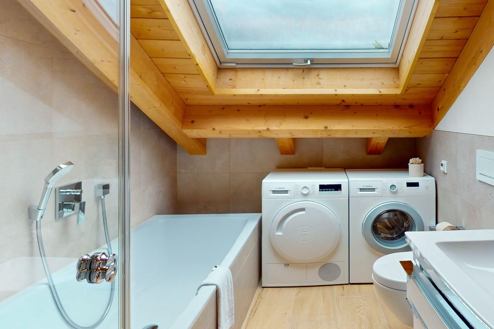 Badezimmer mit Badewanne, Waschmaschine und Trockner unter Holzdecke mit Dachfenster.